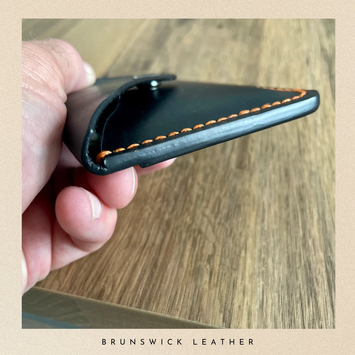 Hand holding a black leather cardholder with orange stitching on a wooden surface, branded 'Brunswick Leather'.