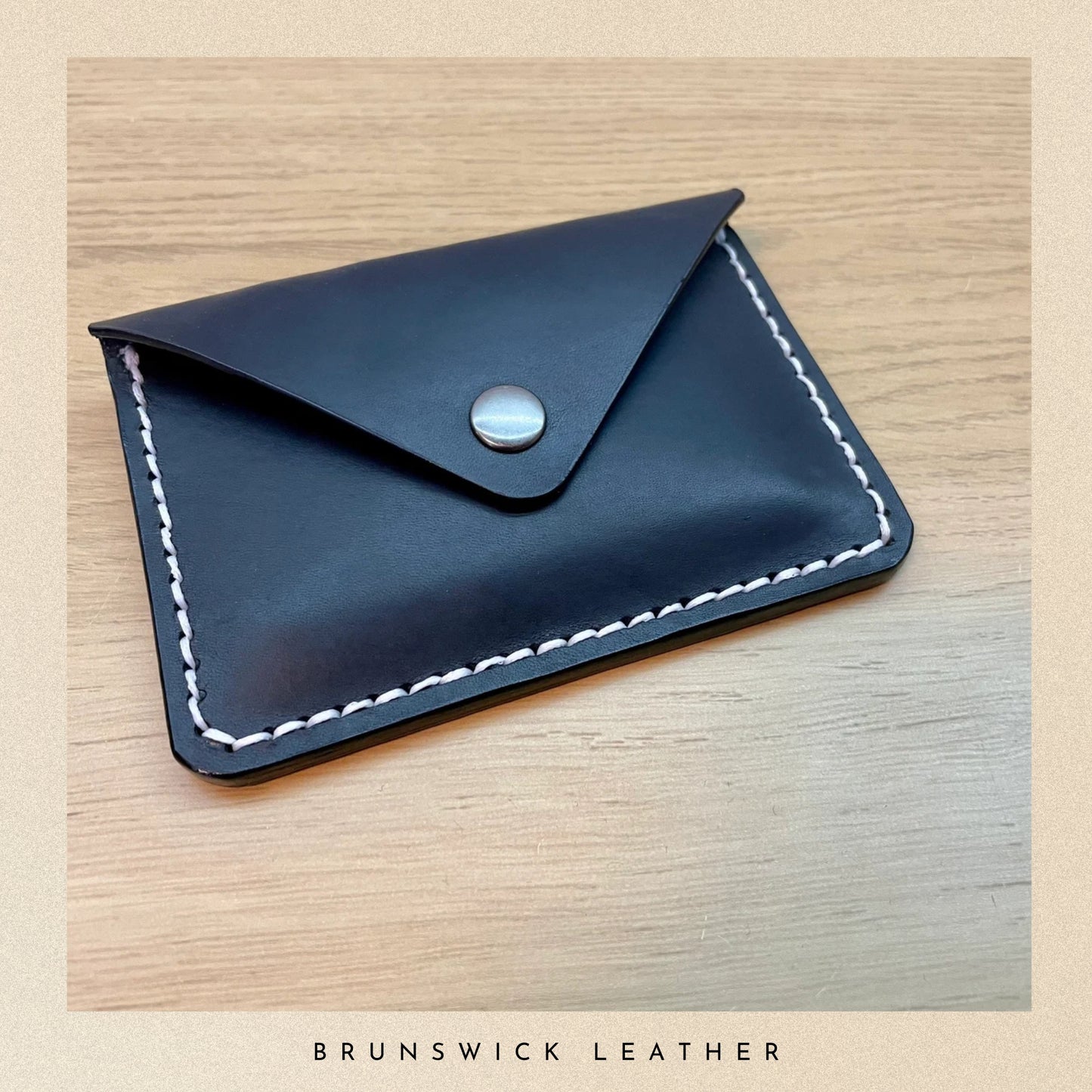 Black leather cardholder with white stitching on a wooden surface, branded 'Brunswick Leather'.