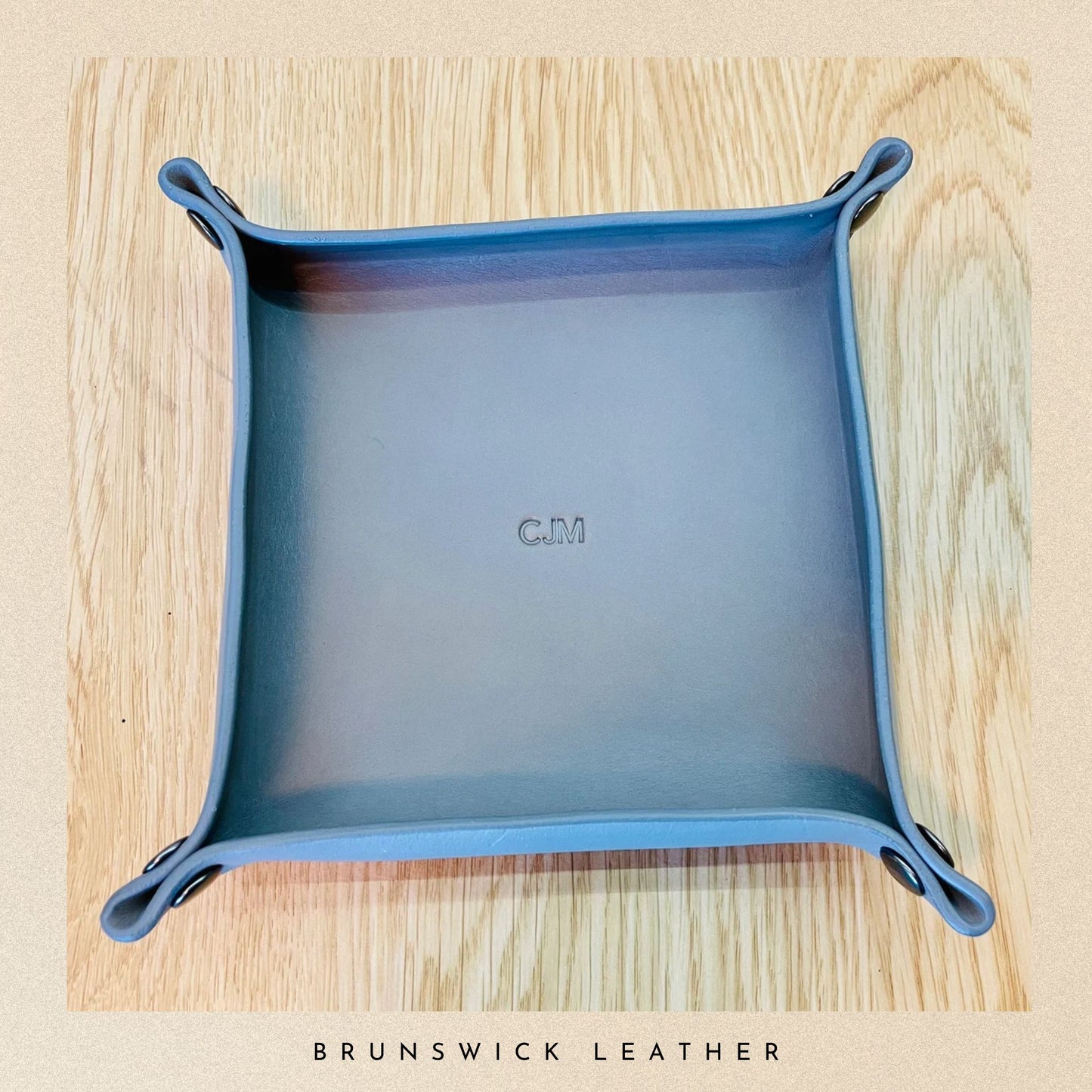 Blue leather tray on a wooden surface with 'C.M' branding, featuring 'Brunswick Leather' text.