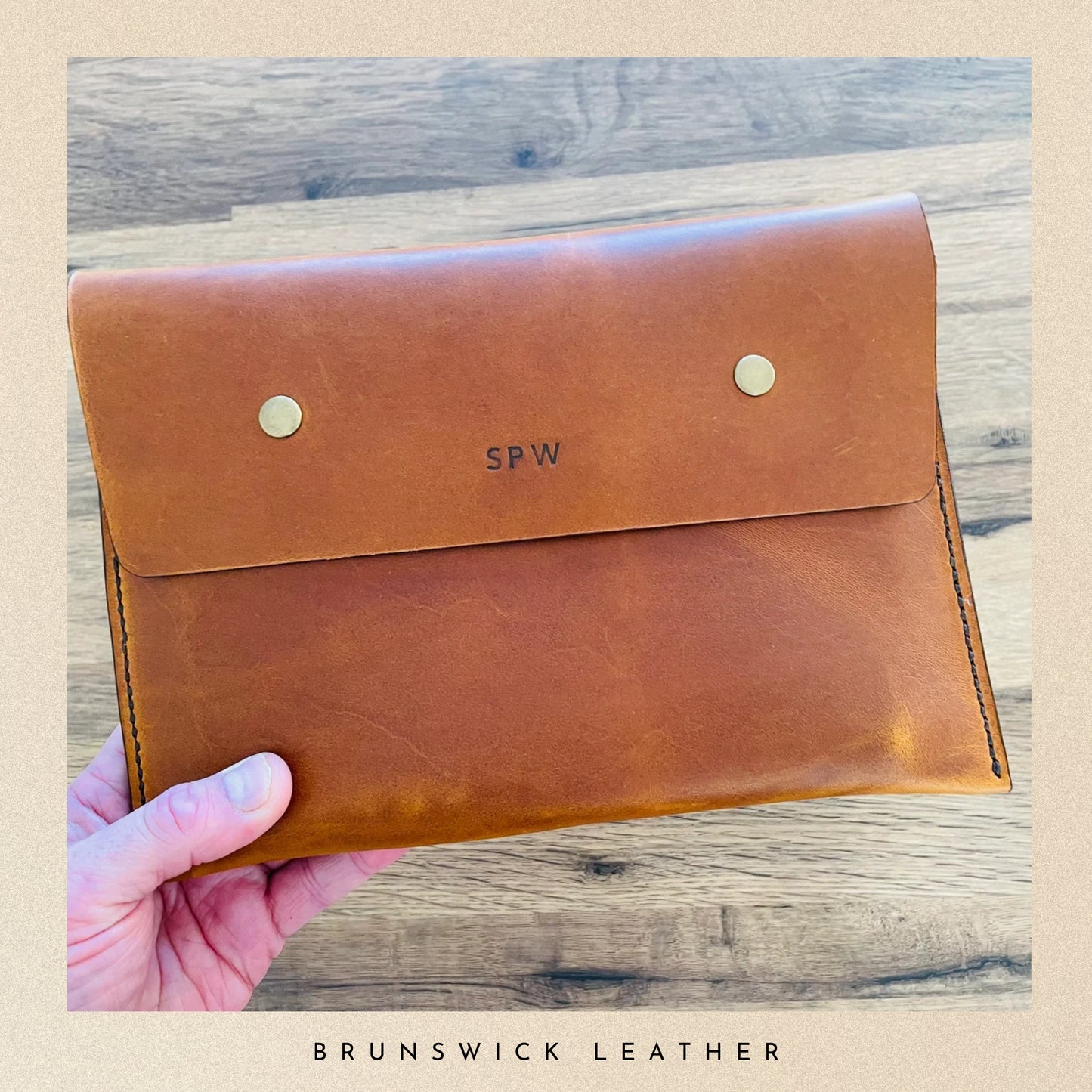 Brown leather clutch held by a hand on a wooden surface with 'Brunswick Leather' branding.