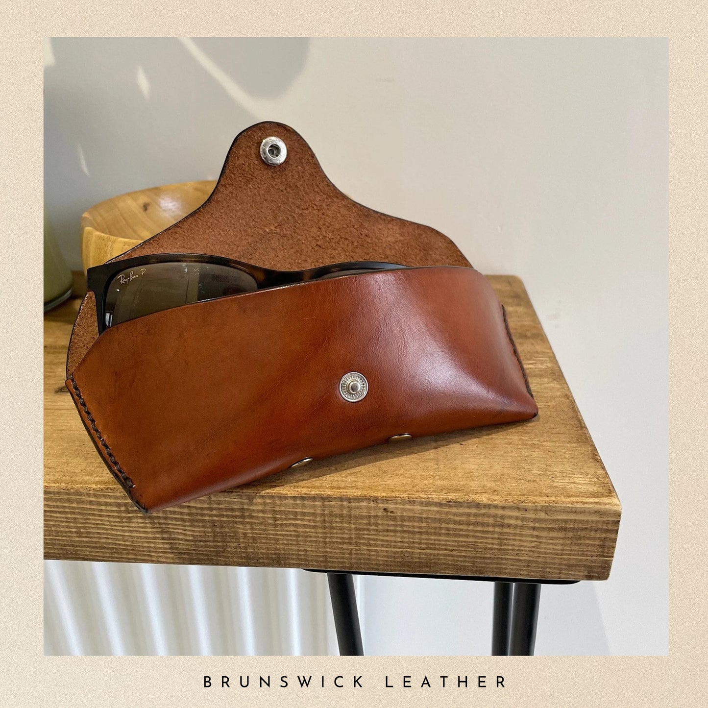 Brown leather sunglasses case on a wooden surface with 'Brunswick Leather' branding.