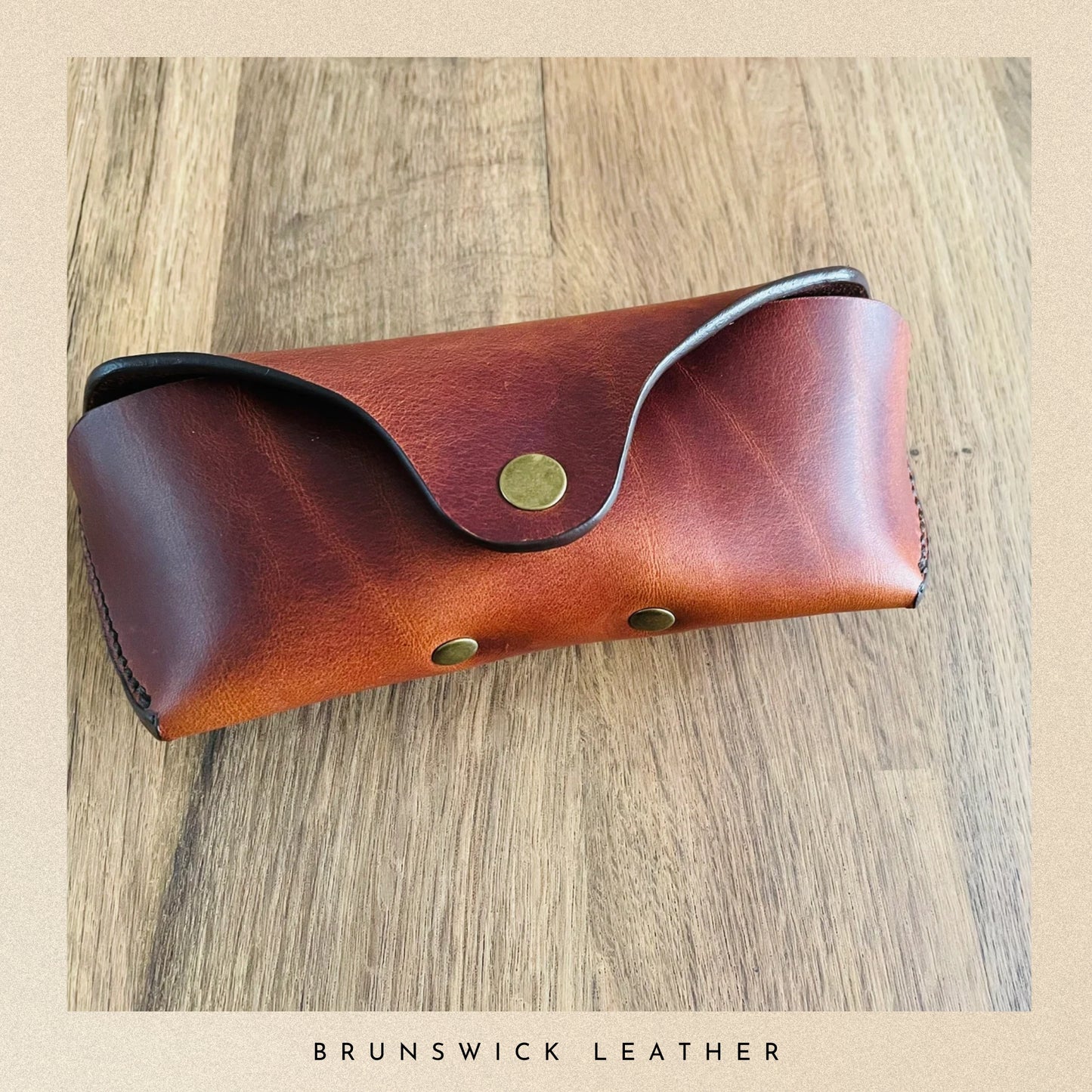 Brown leather sunglasses case on a wooden surface with 'Brunswick Leather' branding.