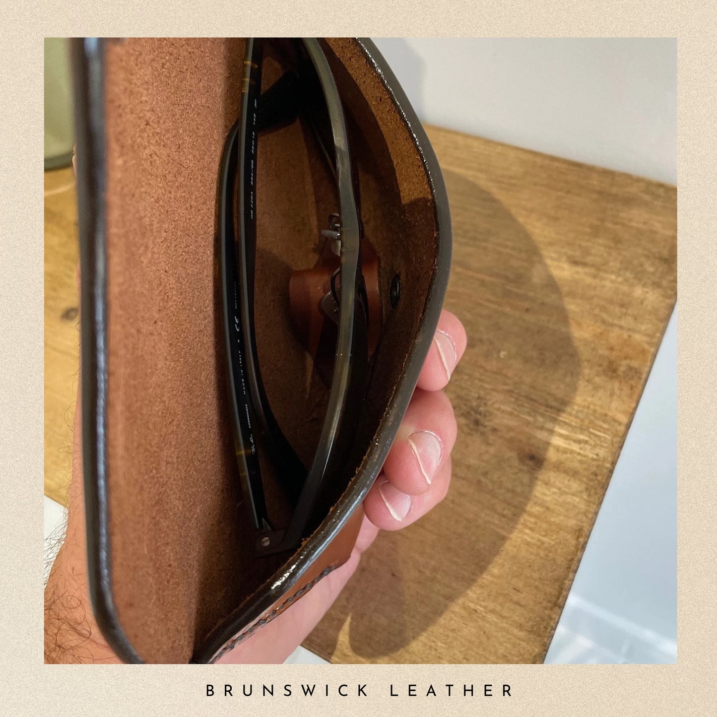 Person holding sunglasses with 'Brunswick Leather' branding on a wooden surface
