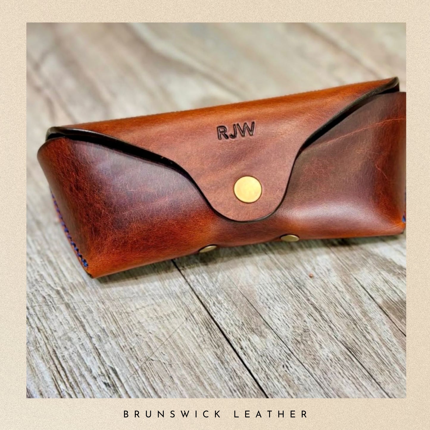 Brown leather glasses case with gold button and 'RJW' engraving on a wooden surface, branded 'Brunswick Leather'.