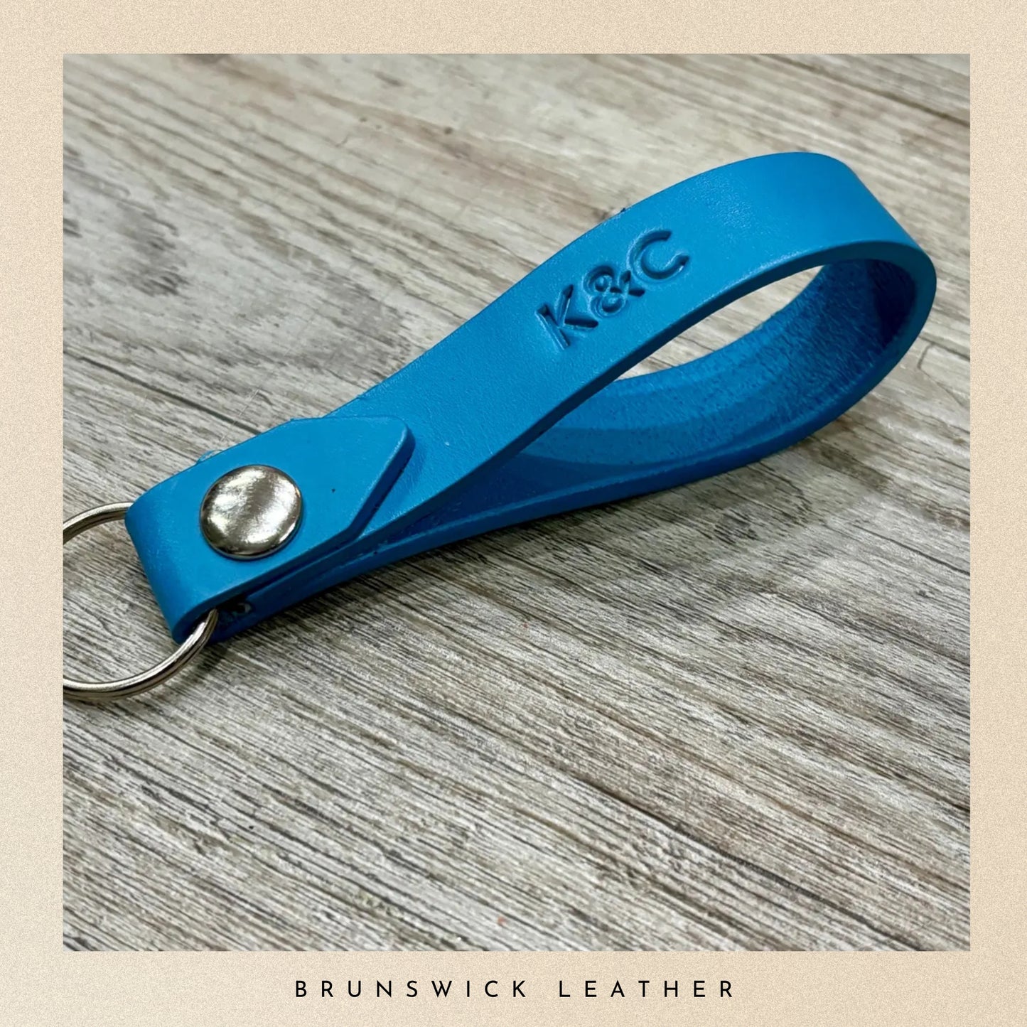 Blue leather keychain with 'K&C' engraving on a wooden surface, branded 'Brunswick Leather'.