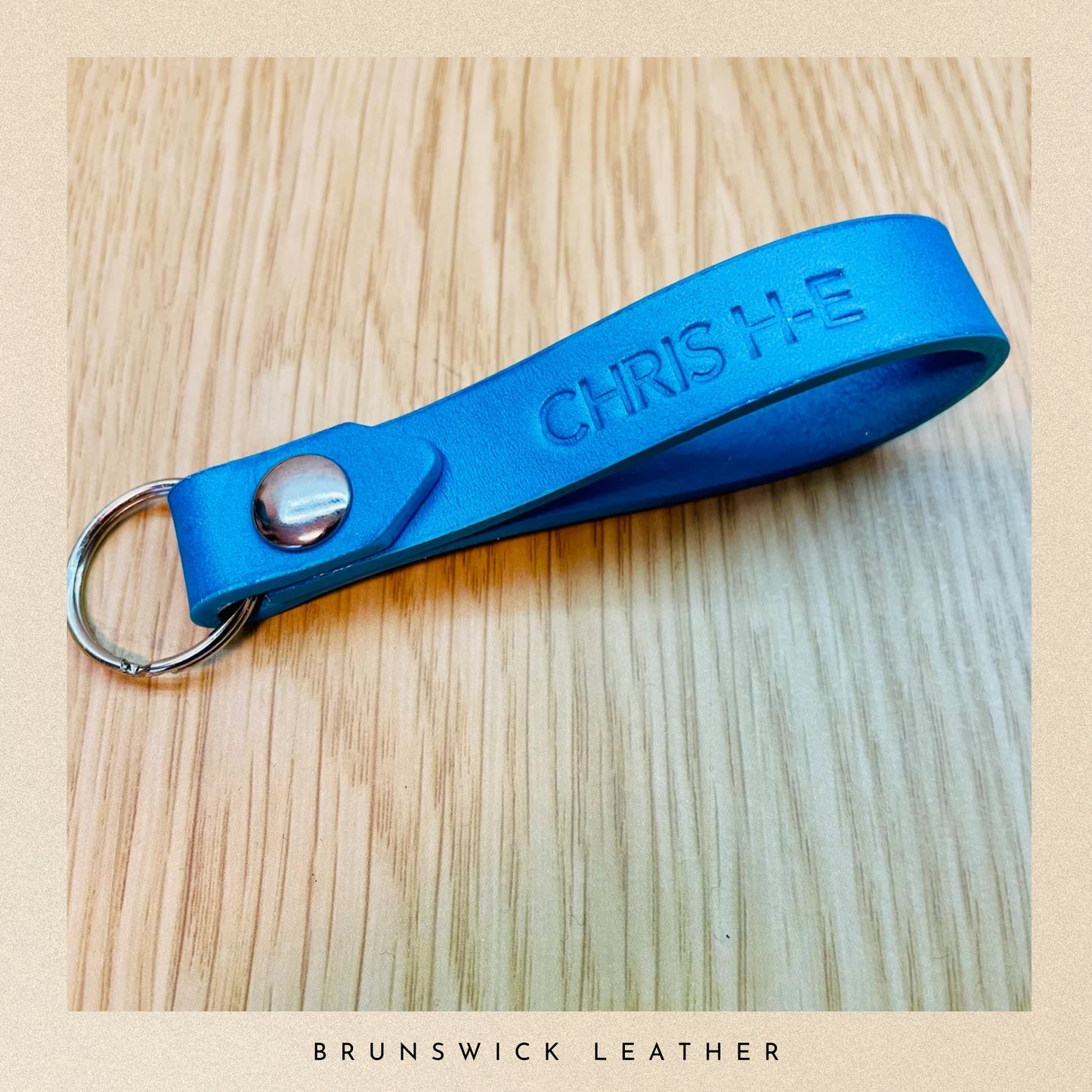 Blue leather keychain with 'CHRIS HE' engraving on a wooden surface, branded 'Brunswick Leather'.
