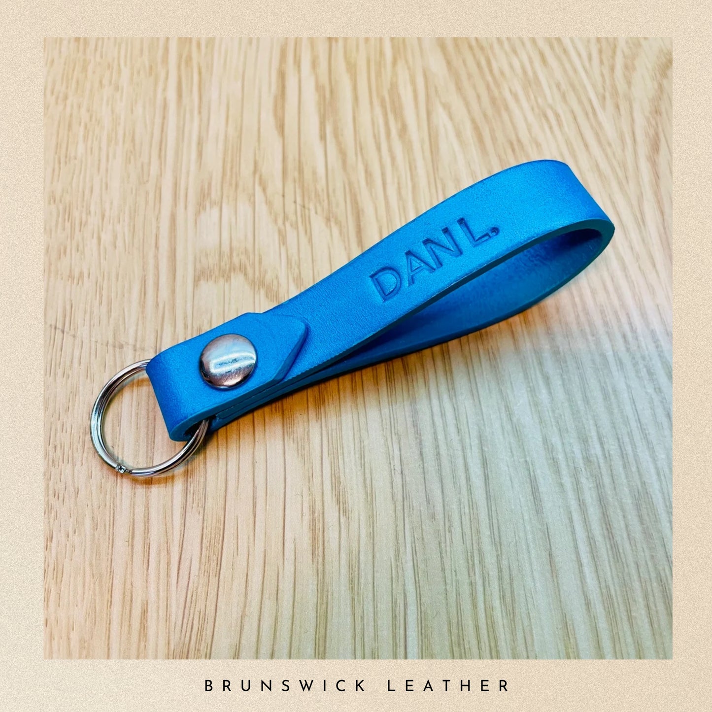 Blue leather keychain with 'DANL' engraving on a wooden surface, branded 'Brunswick Leather'.