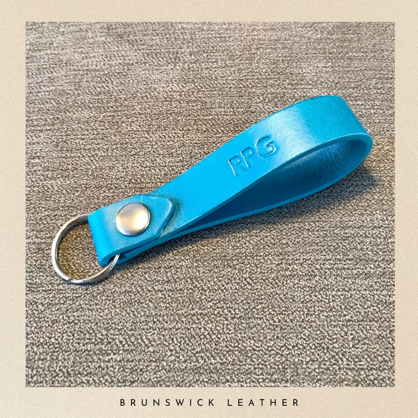Blue leather keychain with a metal ring on a textured beige background, branded 'Brunswick Leather'.