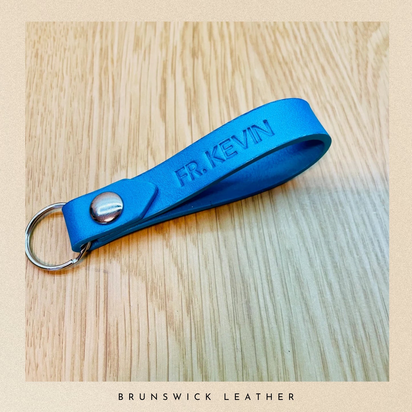 Blue leather keychain with 'FR. KEVIN' engraving on a wooden surface, branded 'Brunswick Leather'.