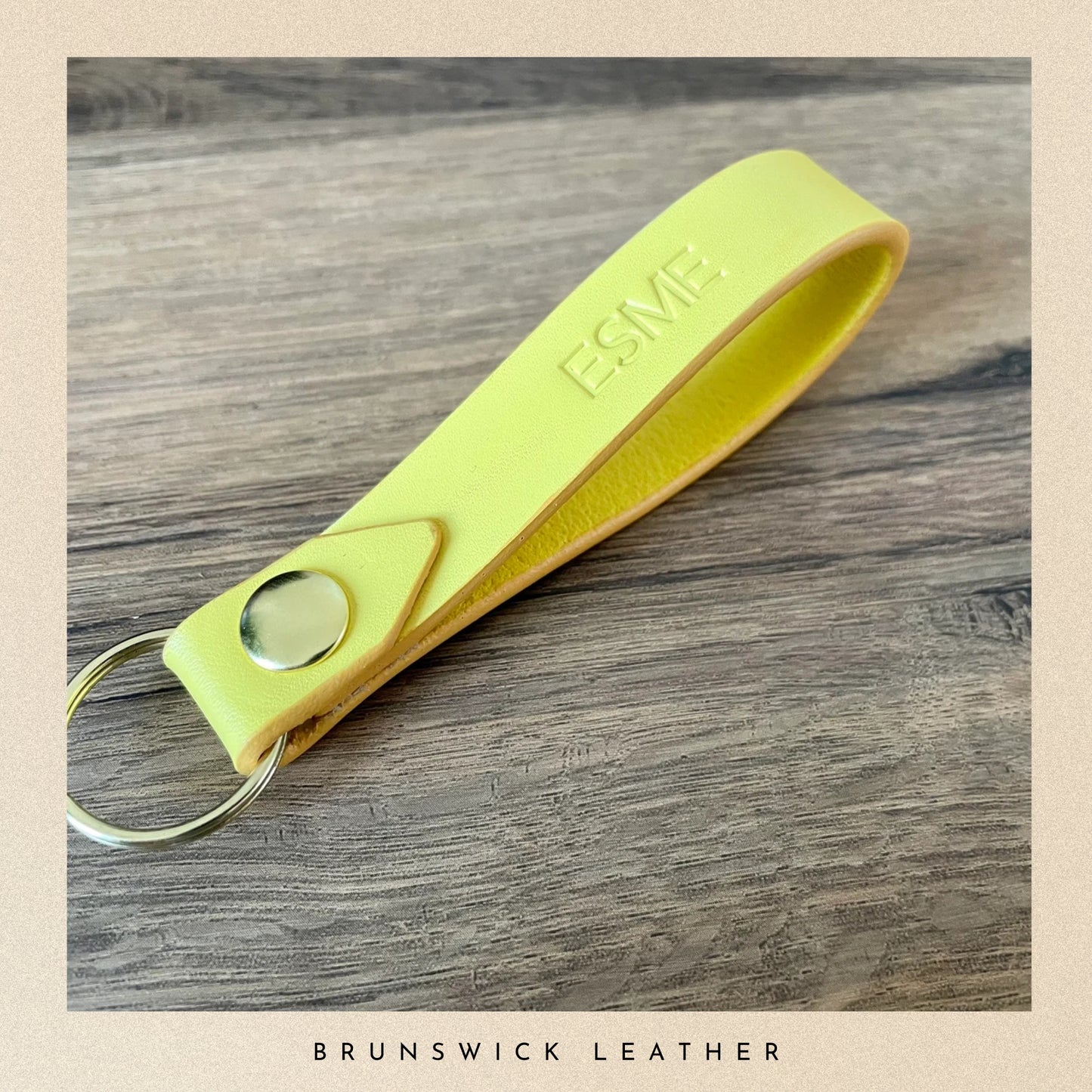 Yellow leather key fob with 'ESME' engraving on a wooden surface, branded 'Brunswick Leather'.