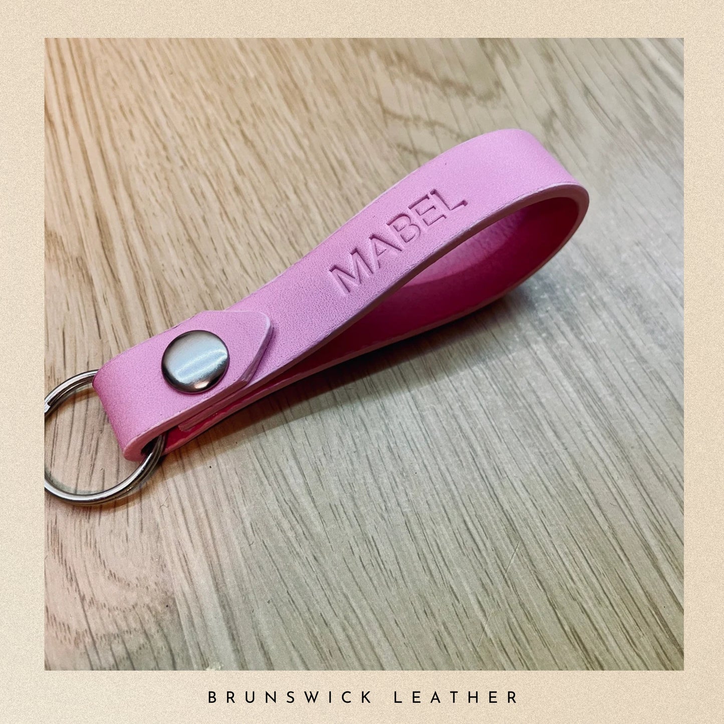 Pink leather keychain with 'MABEL' engraving on a wooden surface, branded 'Brunswick Leather'.