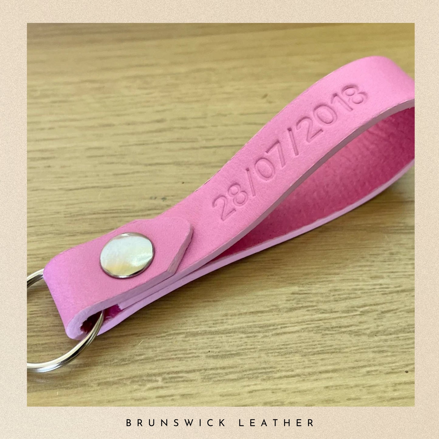 Pink leather keychain with embossed date on a wooden surface, branded 'Brunswick Leather'.