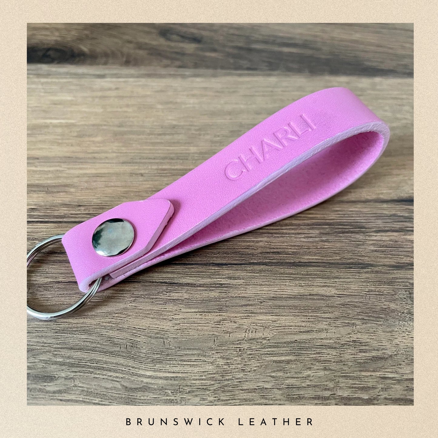 Pink leather keychain with 'CHARM' engraving on a wooden surface, branded 'Brunswick Leather'.