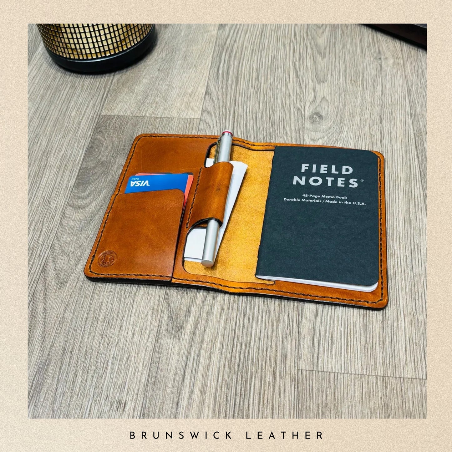 Brown leather wallet with Field Notes notebook and pen on a wooden surface, branded 'Brunswick Leather'.