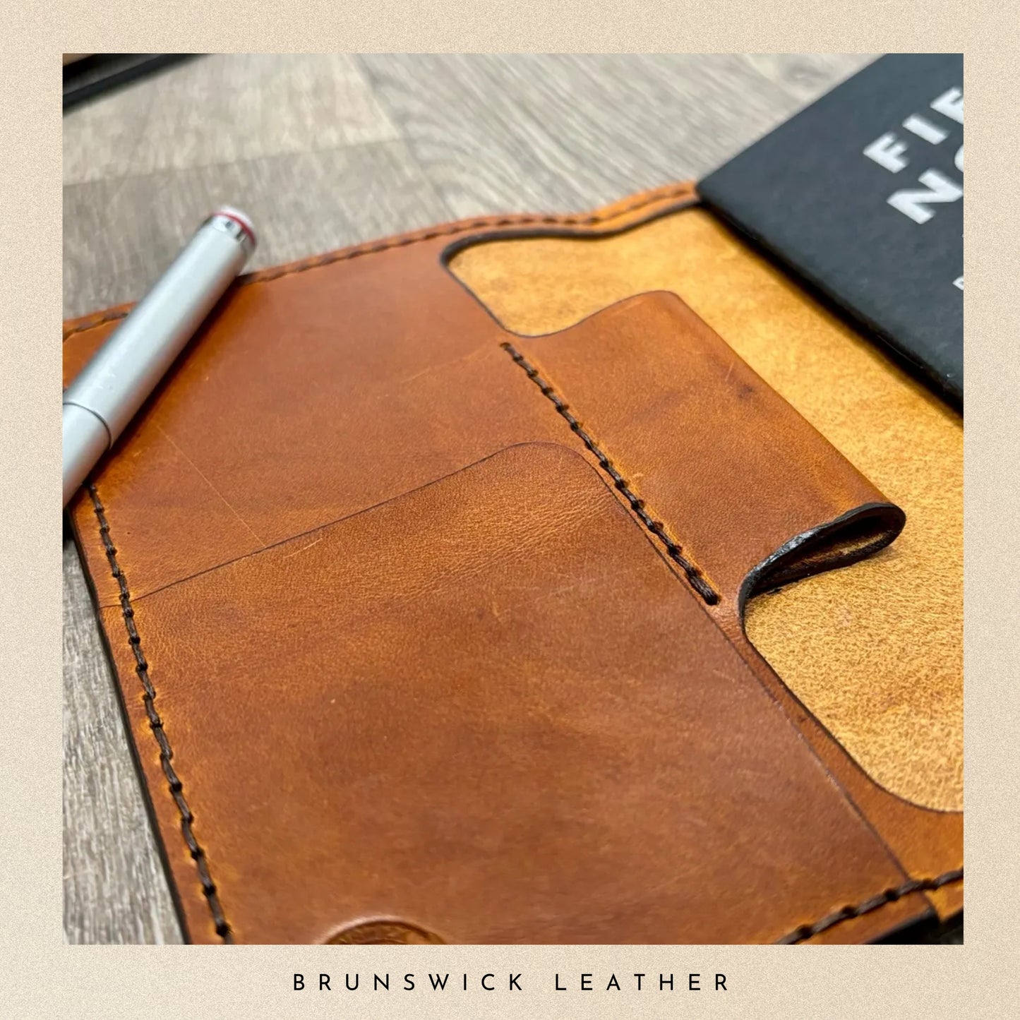 Brown leather notebook with a pen on a wooden surface, branded 'Brunswick Leather'.