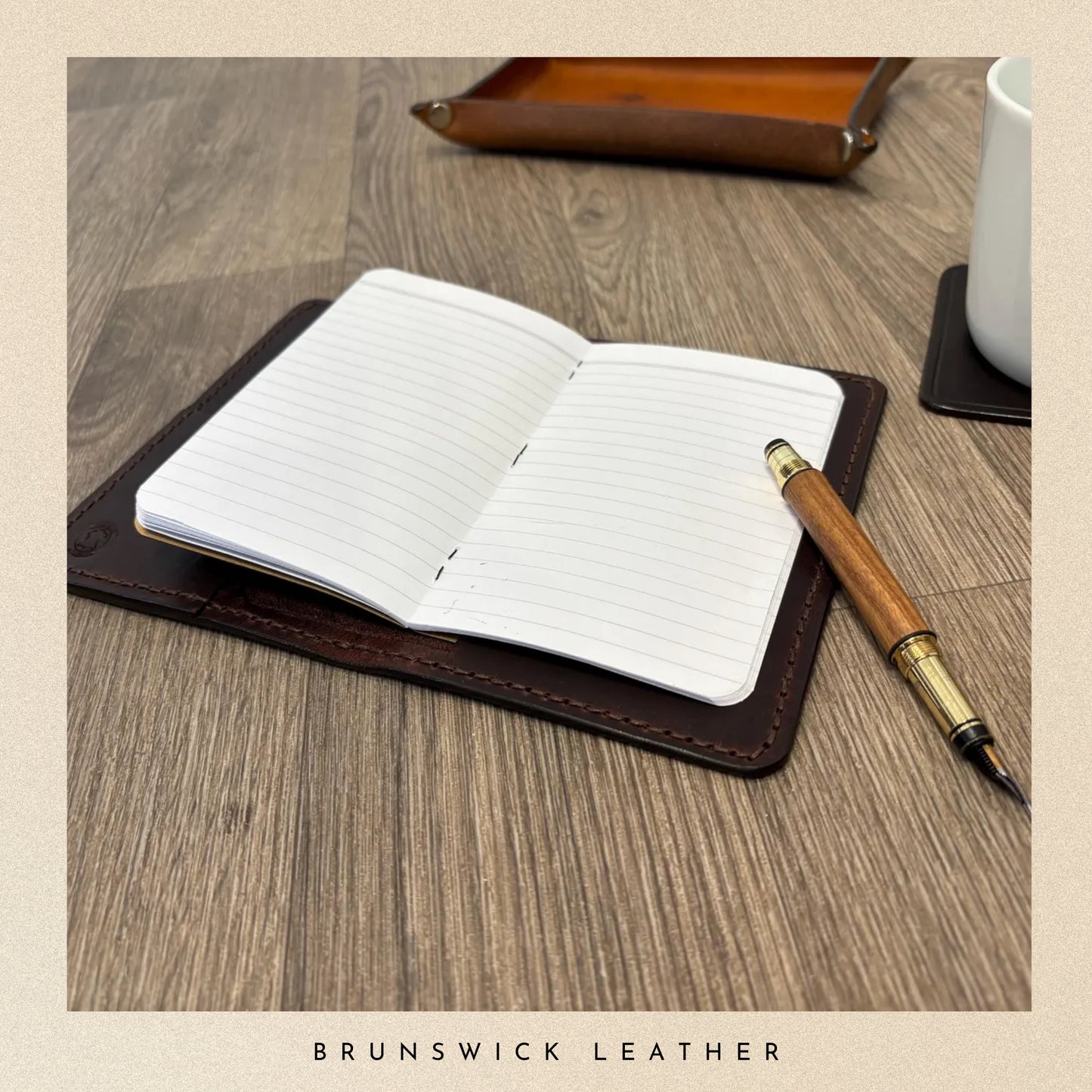 Open notebook with lined pages on a leather stand, accompanied by a wooden pen, on a wooden surface with 'Brunswick Leather' branding.
