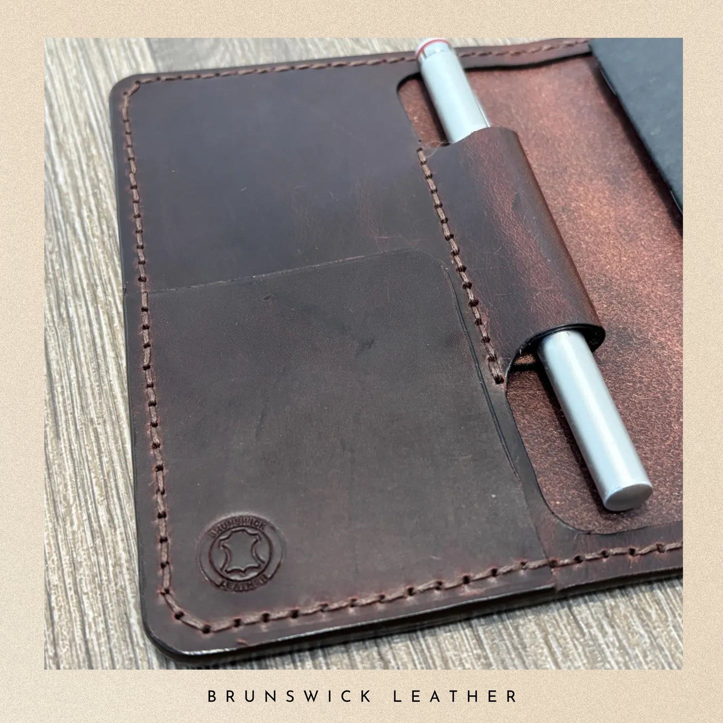 Brown leather notebook cover with a pen on a wooden surface, branded 'Brunswick Leather'.