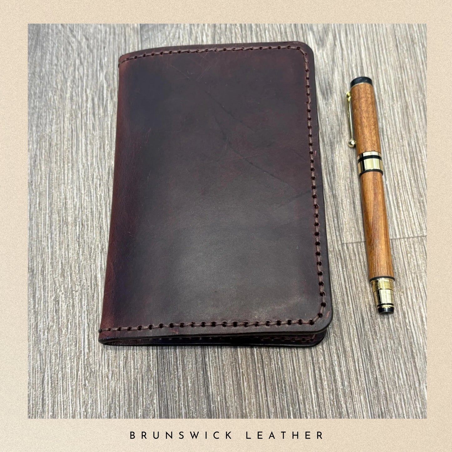 Brown leather notebook and wooden pen on a textured surface with 'Brunswick Leather' branding.