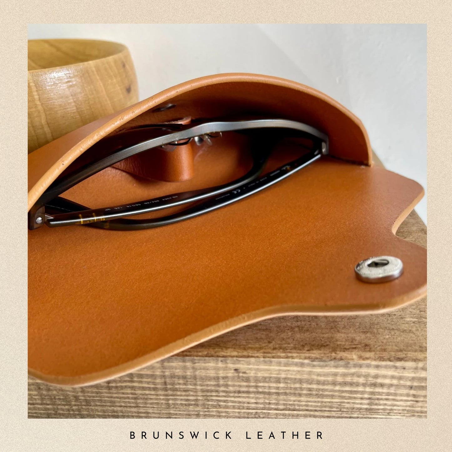 Brown leather case with sunglasses inside, on a wooden surface, branded 'Brunswick Leather'.