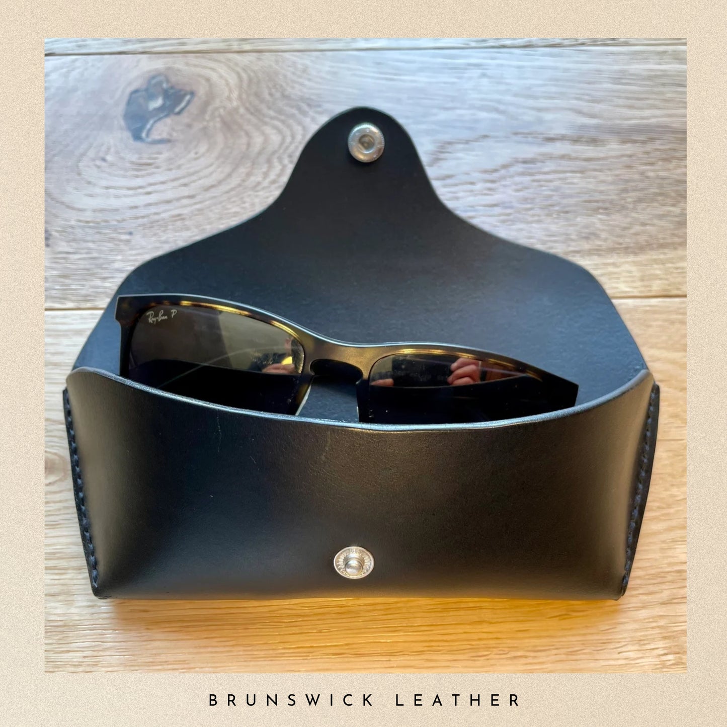 Black leather sunglasses case on a wooden surface with 'Brunswick Leather' branding.