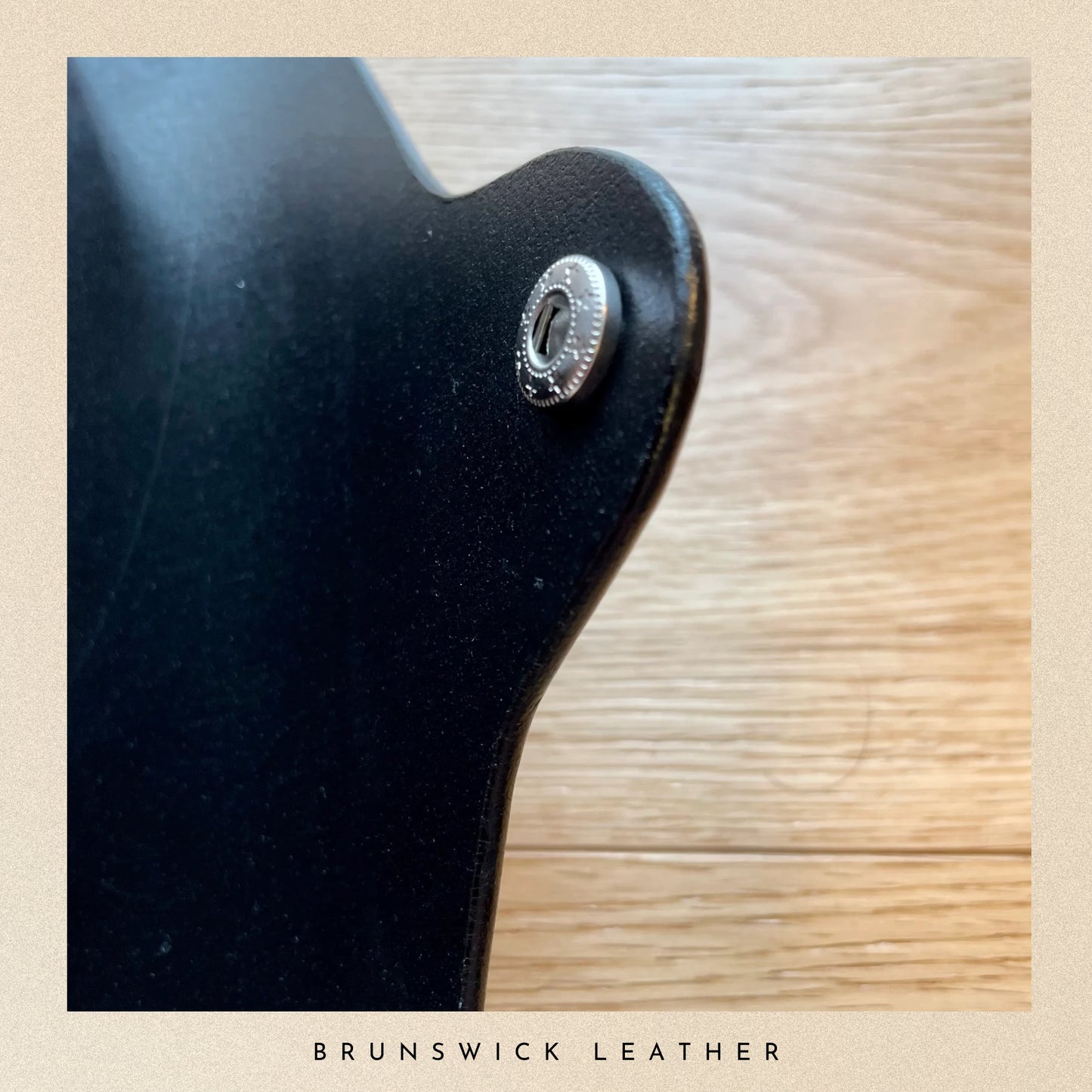 Close-up of a black leather product with a silver button on a wooden surface, branded 'Brunswick Leather'.