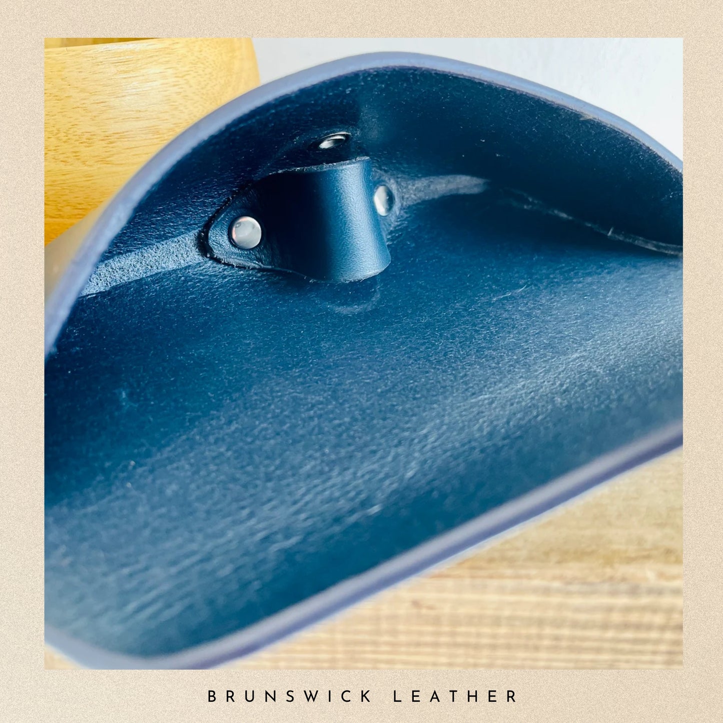 Close-up of a blue leather product with a metal buckle on a beige background, featuring the brand 'Brunswick Leather'.