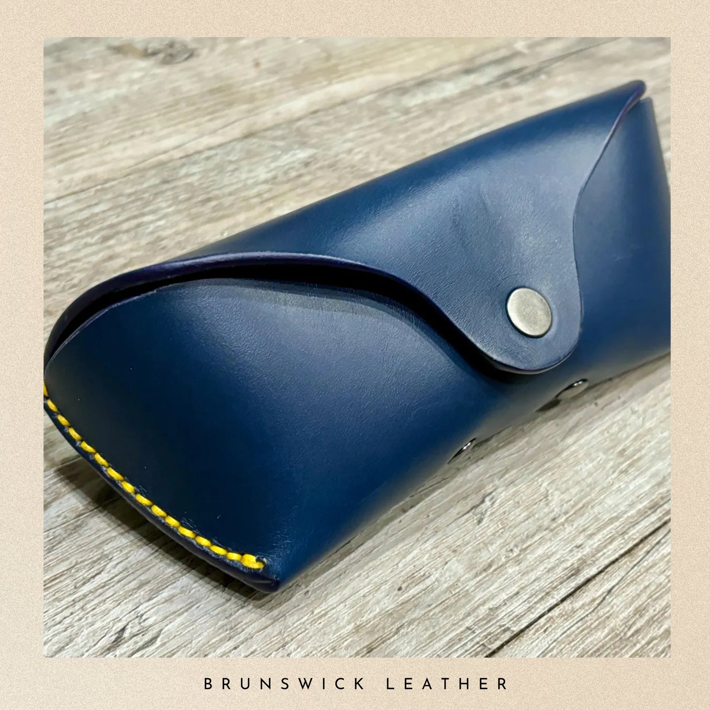 Blue leather case with yellow stitching on a wooden surface, branded 'Brunswick Leather'.