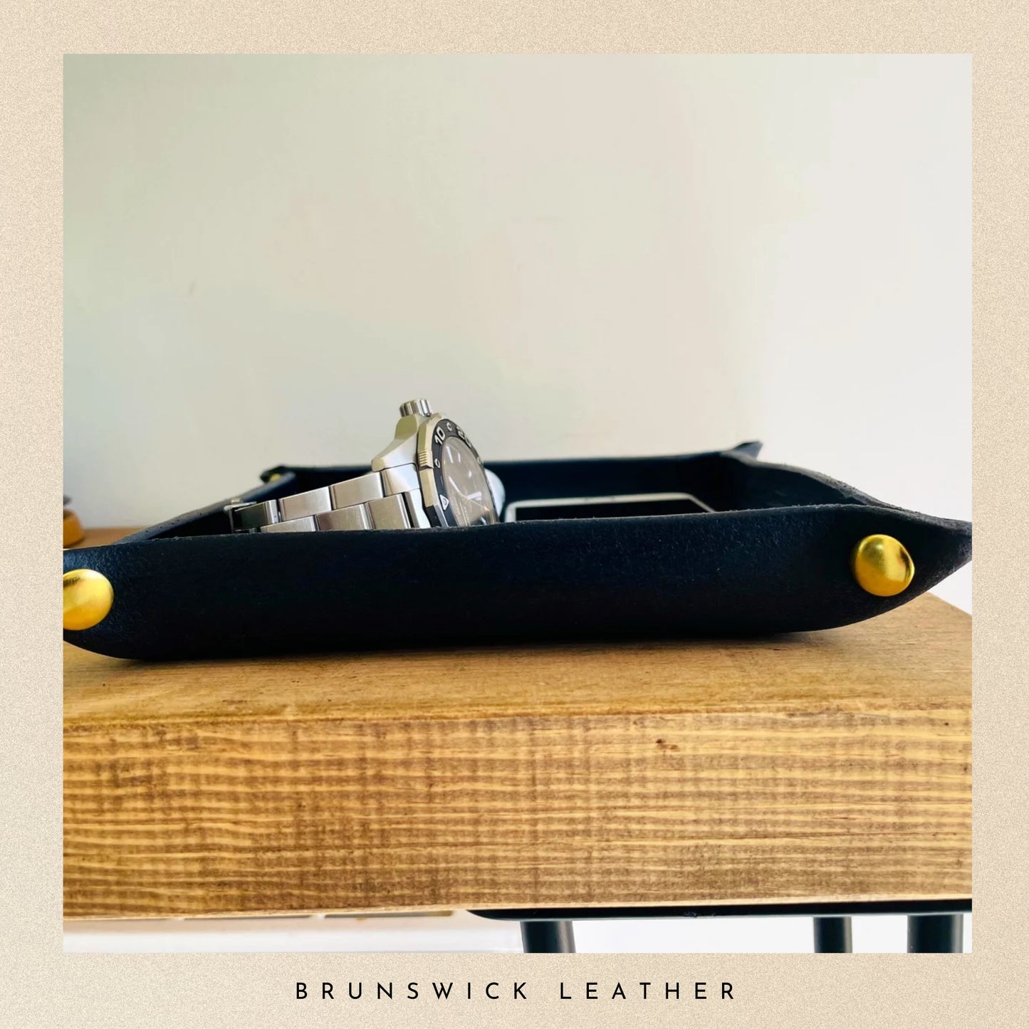 Black leather tray with gold accents holding a watch on a wooden surface, branded 'Brunswick Leather'.