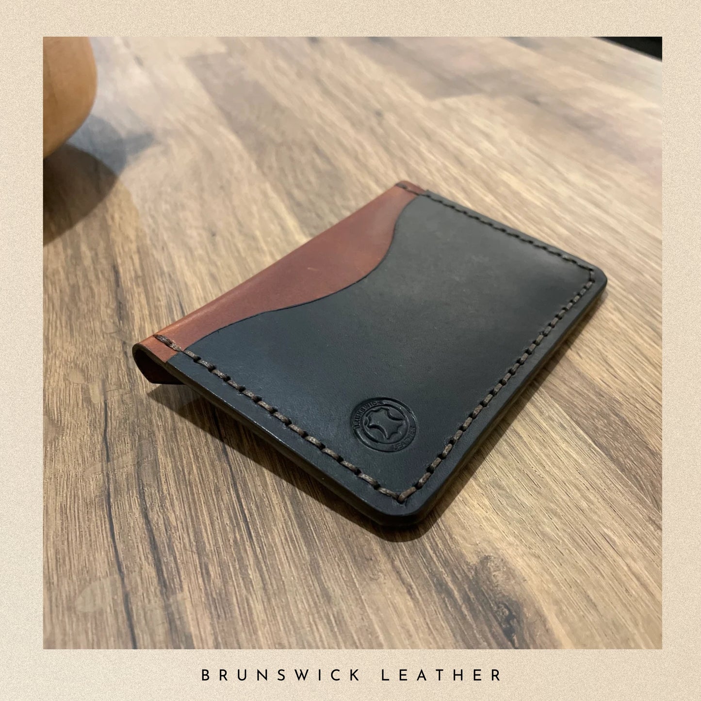 Black leather wallet with brown accent on a wooden surface, branded 'Brunswick Leather'.