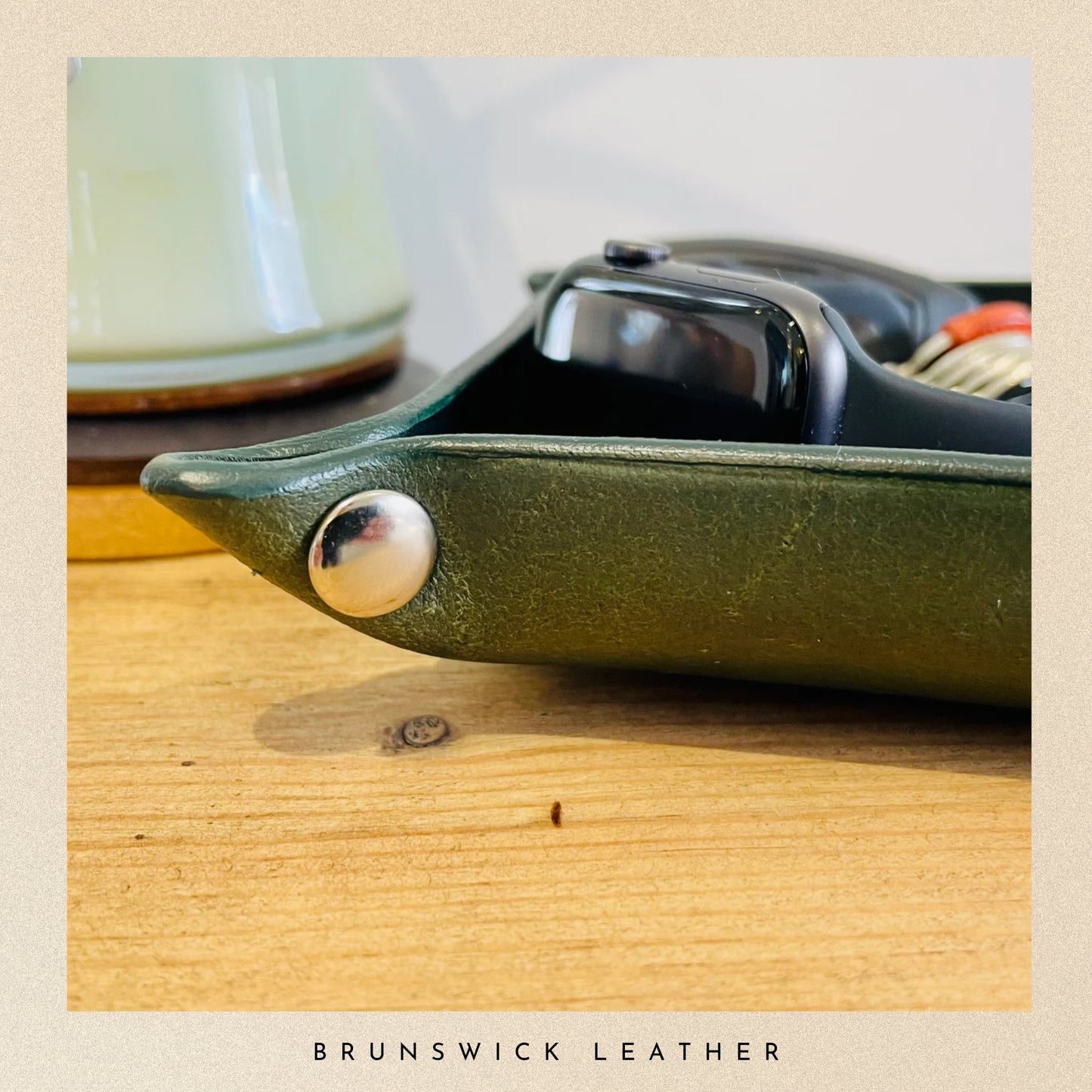 Green leather case with silver button on a wooden surface, featuring 'Brunswick Leather' branding.