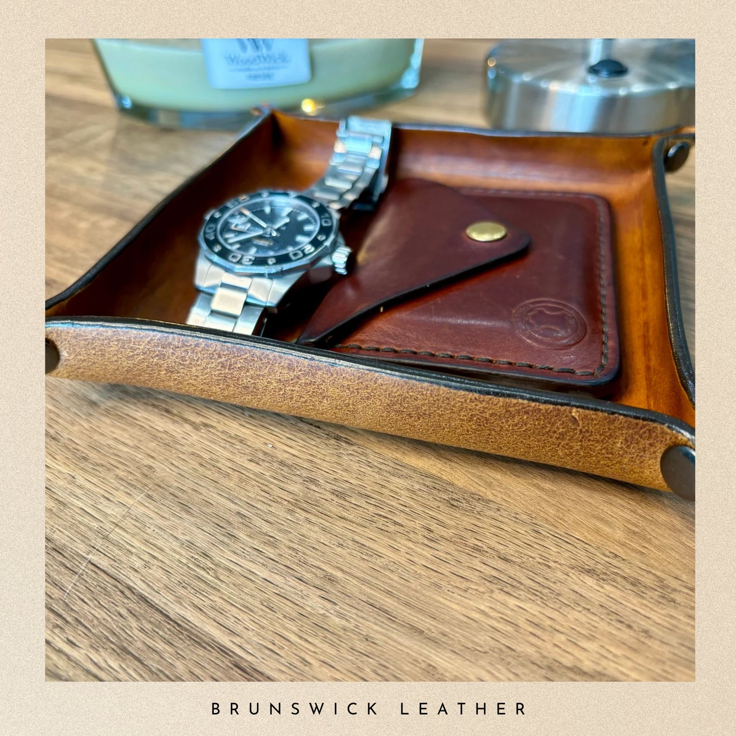 Brown leather wallet with a watch inside on a wooden surface, branded 'Brunswick Leather'.