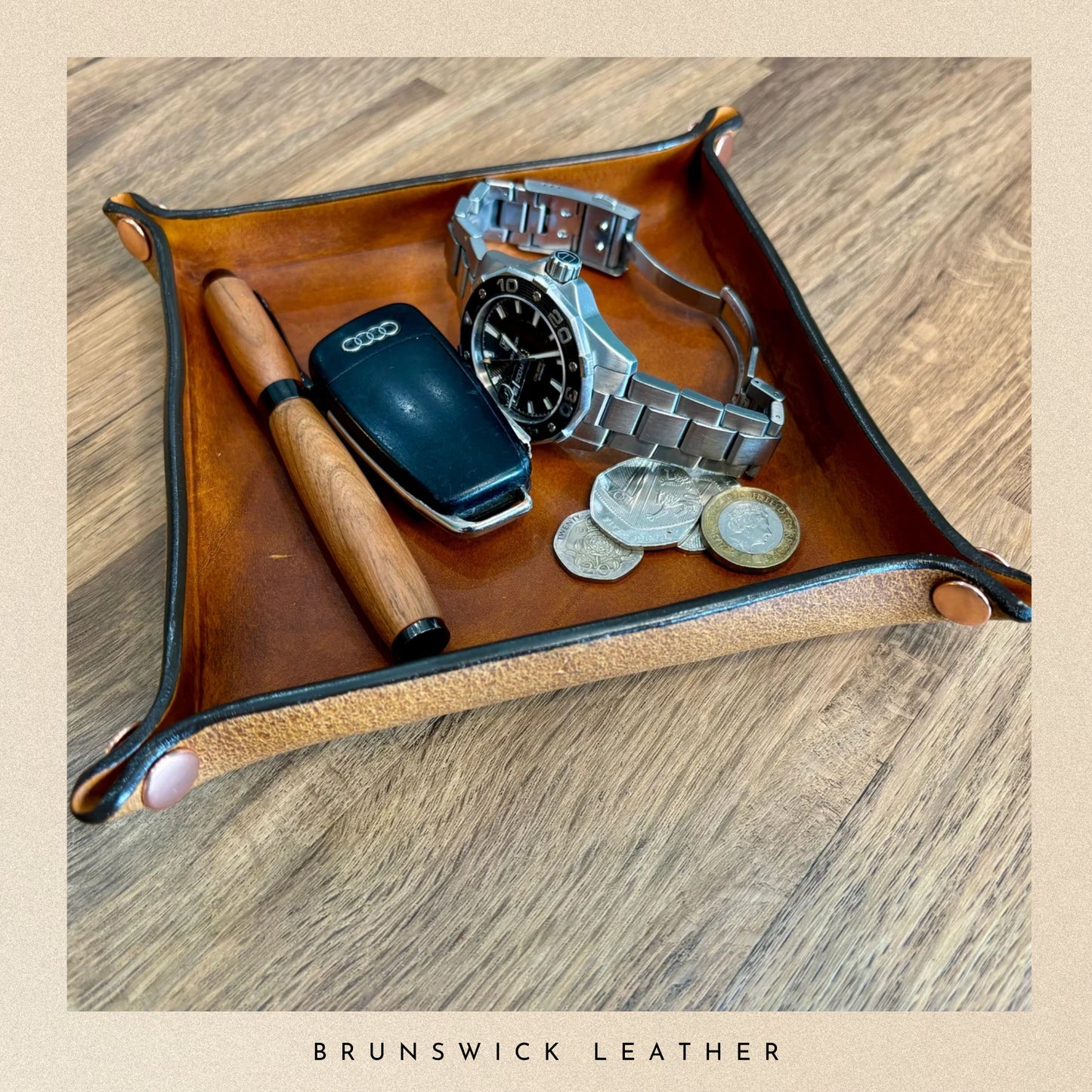 Brown leather tray with a watch, phone, coins, and a pen on a wooden surface, branded 'Brunswick Leather'.