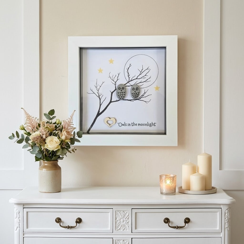A framed wall art piece displayed above a white dresser in a styled room. The artwork shows two grey owls perched on a bare tree branch in front of a large moon, with small gold stars and the phrase 'Owl in the moonlight' near the bottom.