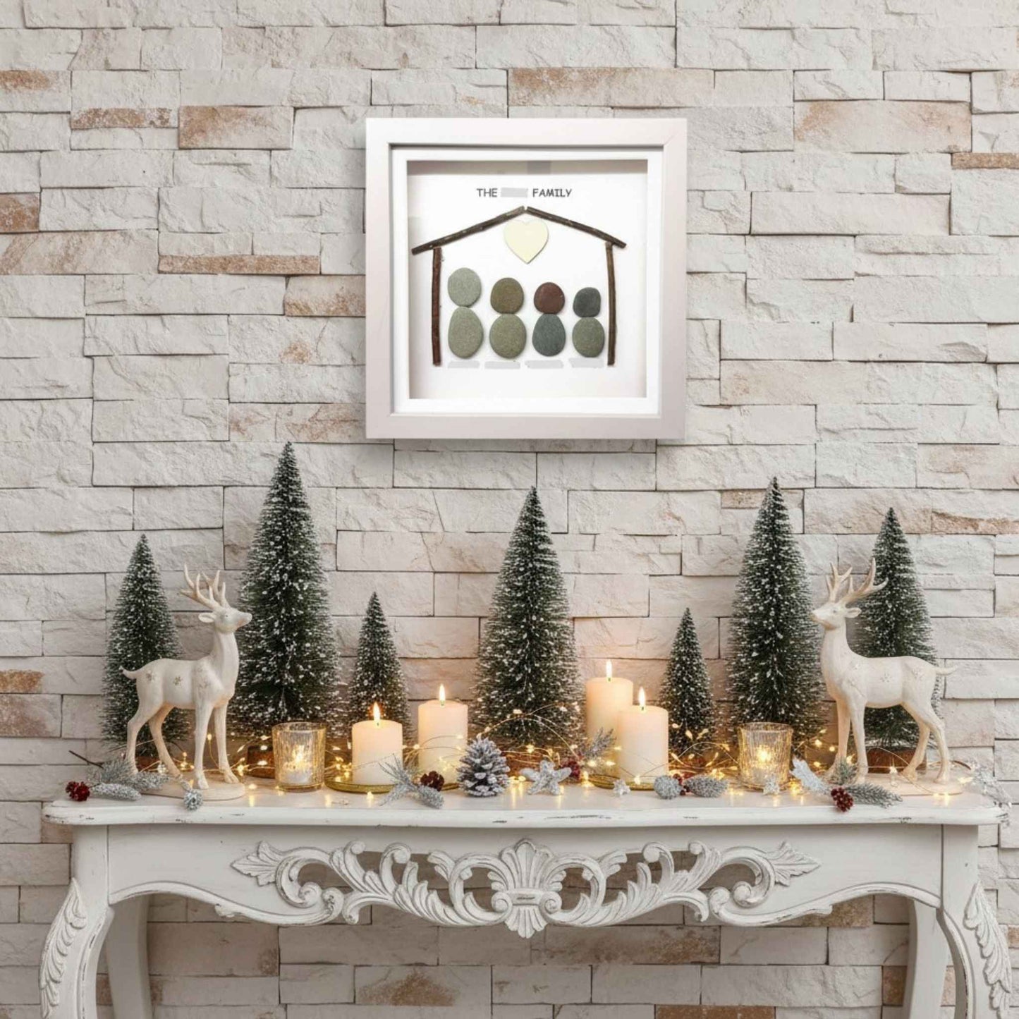 Decorative setup with Christmas trees, candles, and deer figurines on a mantelpiece against a stone wall. A pretty family pebble art frame is above.