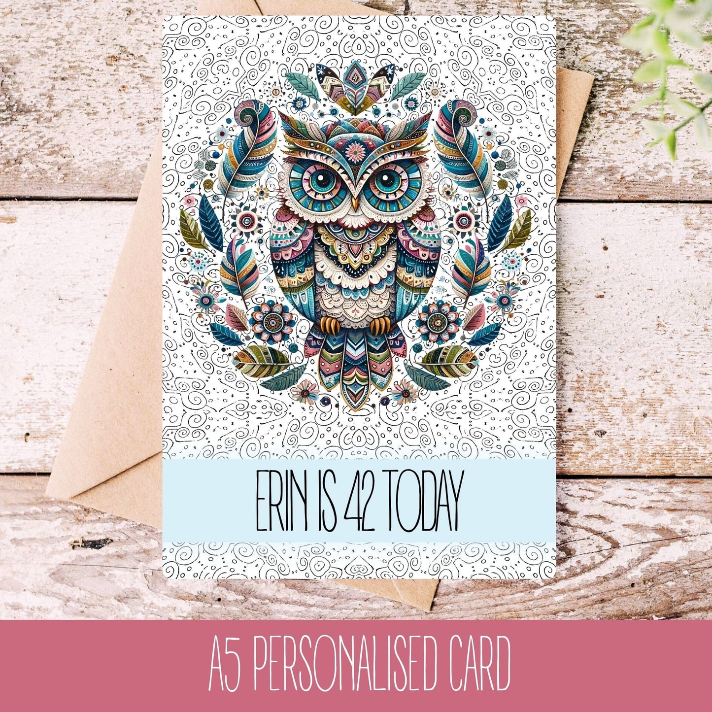 A personalised greeting card with a colorful owl illustration, featuring text that reads 'Erin is 42 today'. The card is placed on a wooden surface with a white envelope beside it.