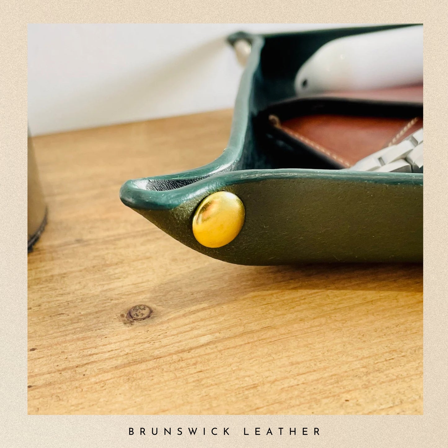 Green leather product with a gold button on a wooden surface, branded 'Brunswick Leather'.