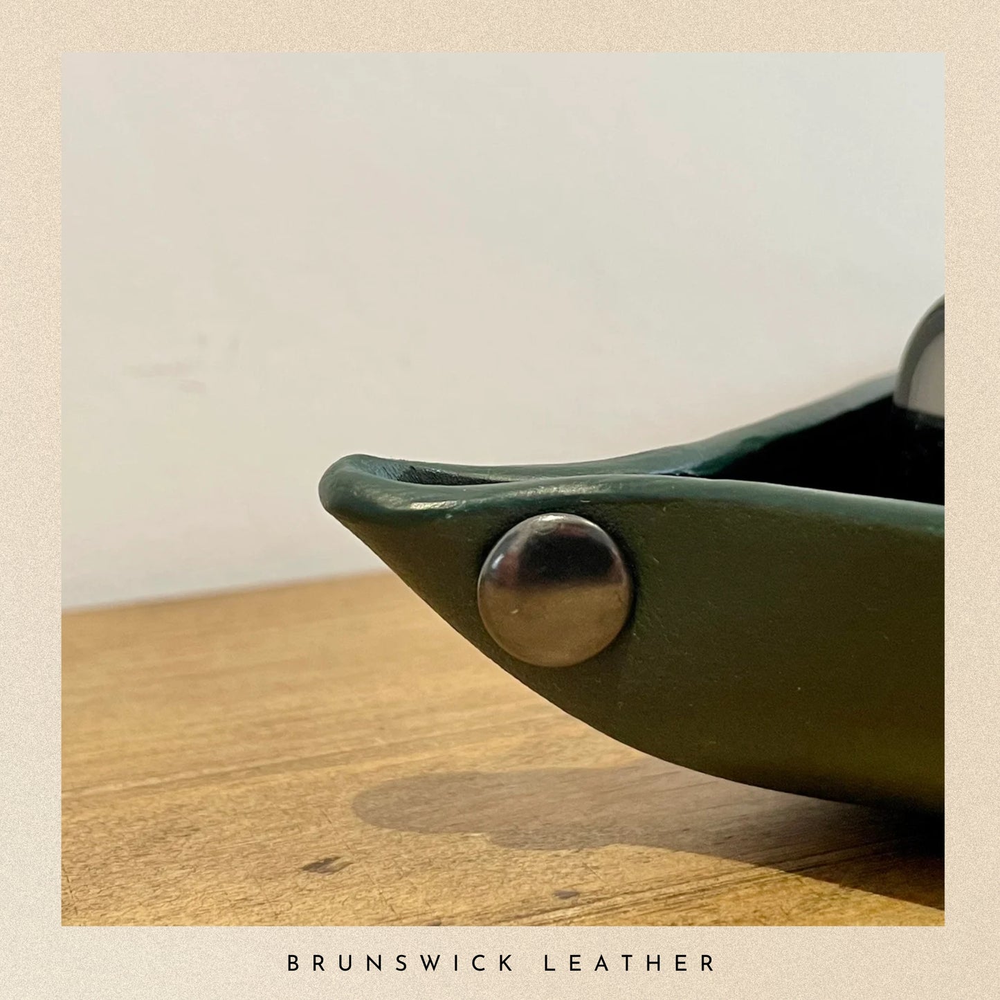 Close-up of a green leather product with a silver button on a wooden surface, branded 'Brunswick Leather'.