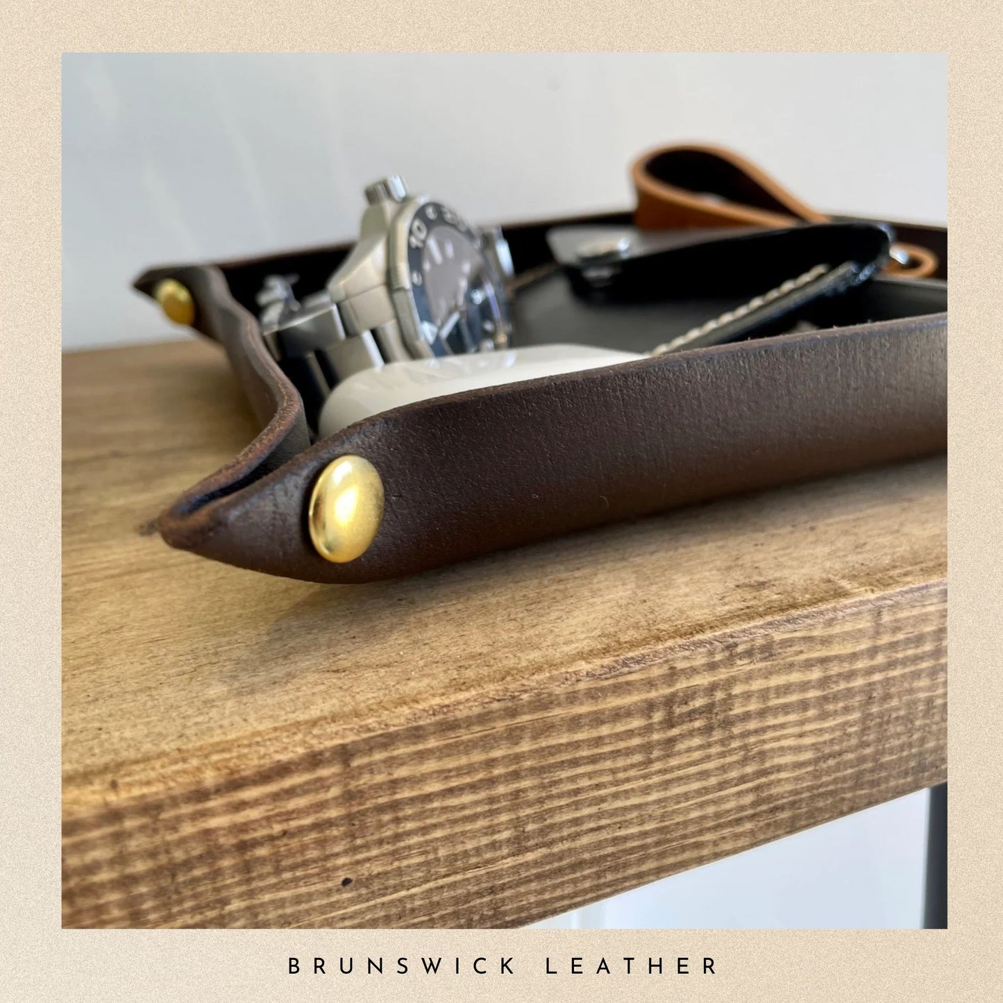 Brown leather tray with gold accents on a wooden surface, featuring the brand 'Brunswick Leather'.