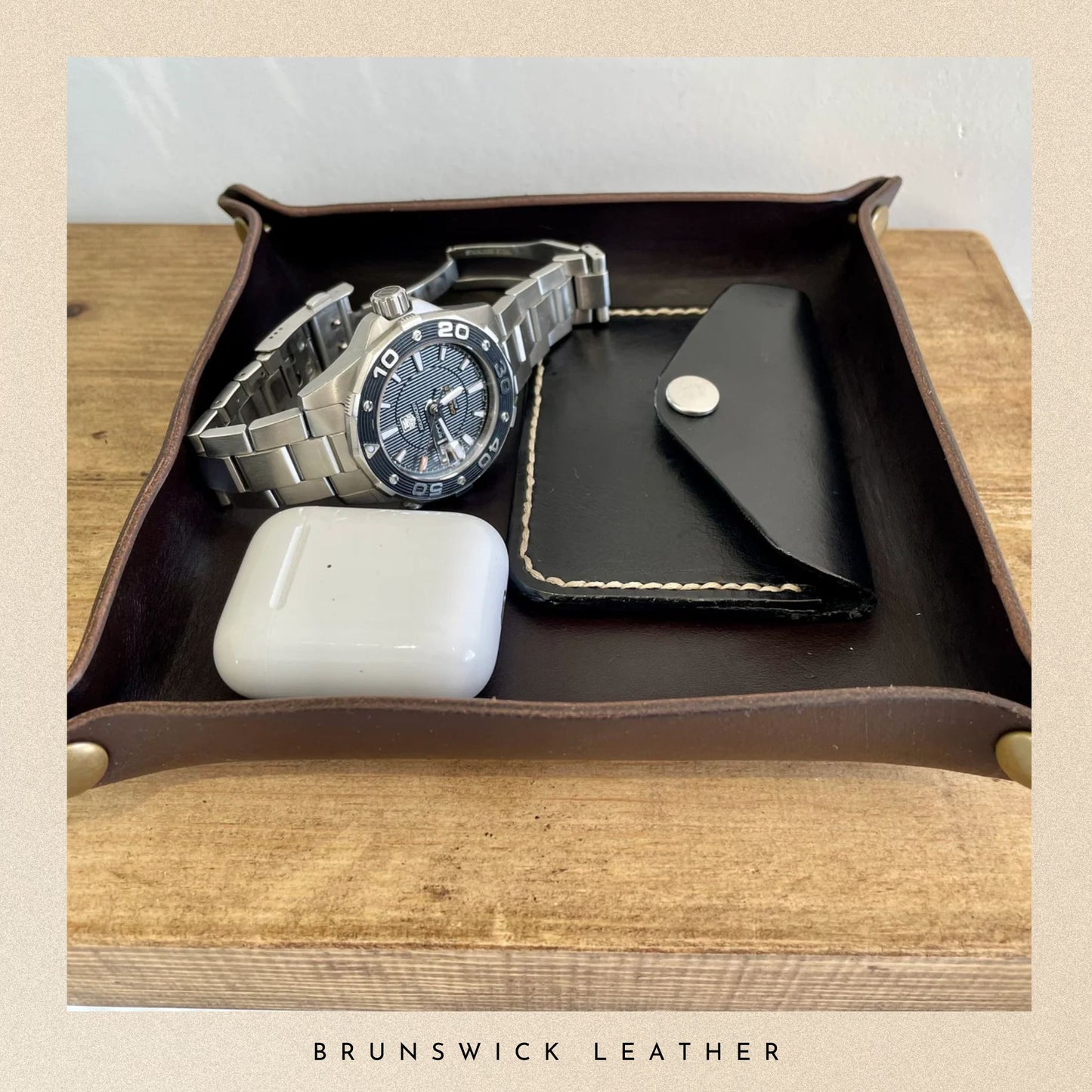 Brown leather tray with a watch, phone case, and white object on a wooden surface, branded 'Brunswick Leather'.