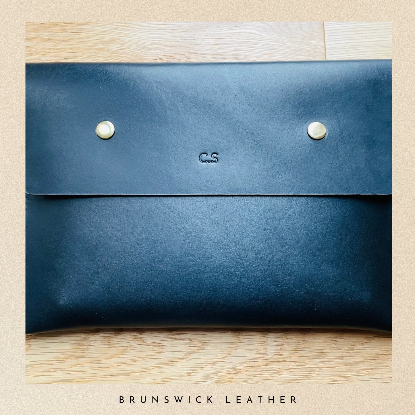 Blue leather clutch with 'CS' initials on a wooden surface, branded 'Brunswick Leather'.