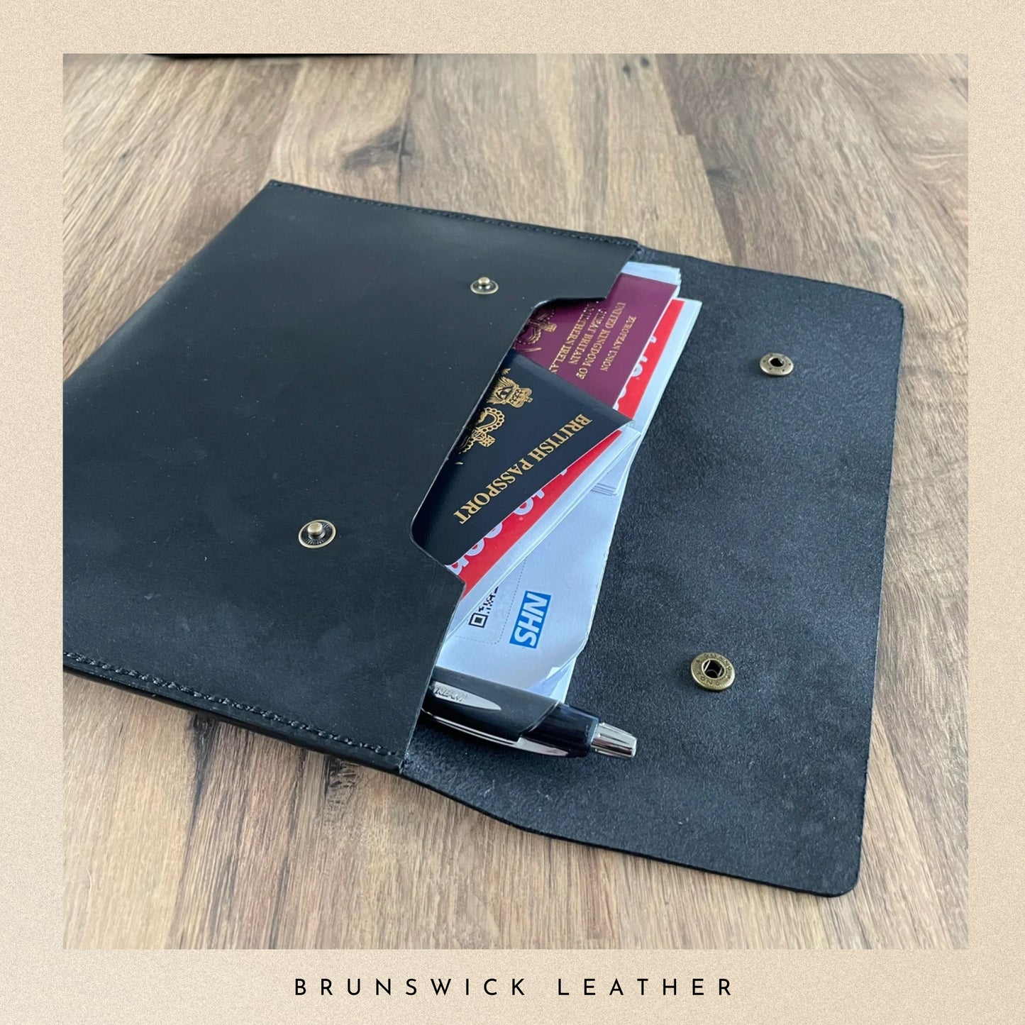 Black leather wallet with a pen and passport holder on a wooden surface, branded 'Brunswick Leather'.