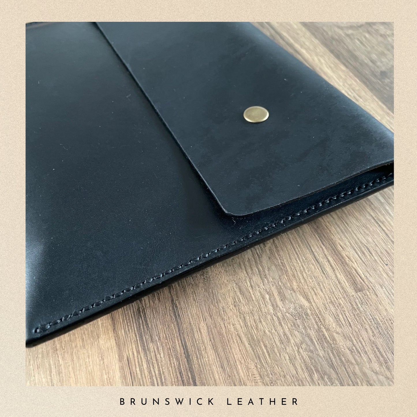 Black leather envelope with a gold button on a wooden surface, branded 'Brunswick Leather'.