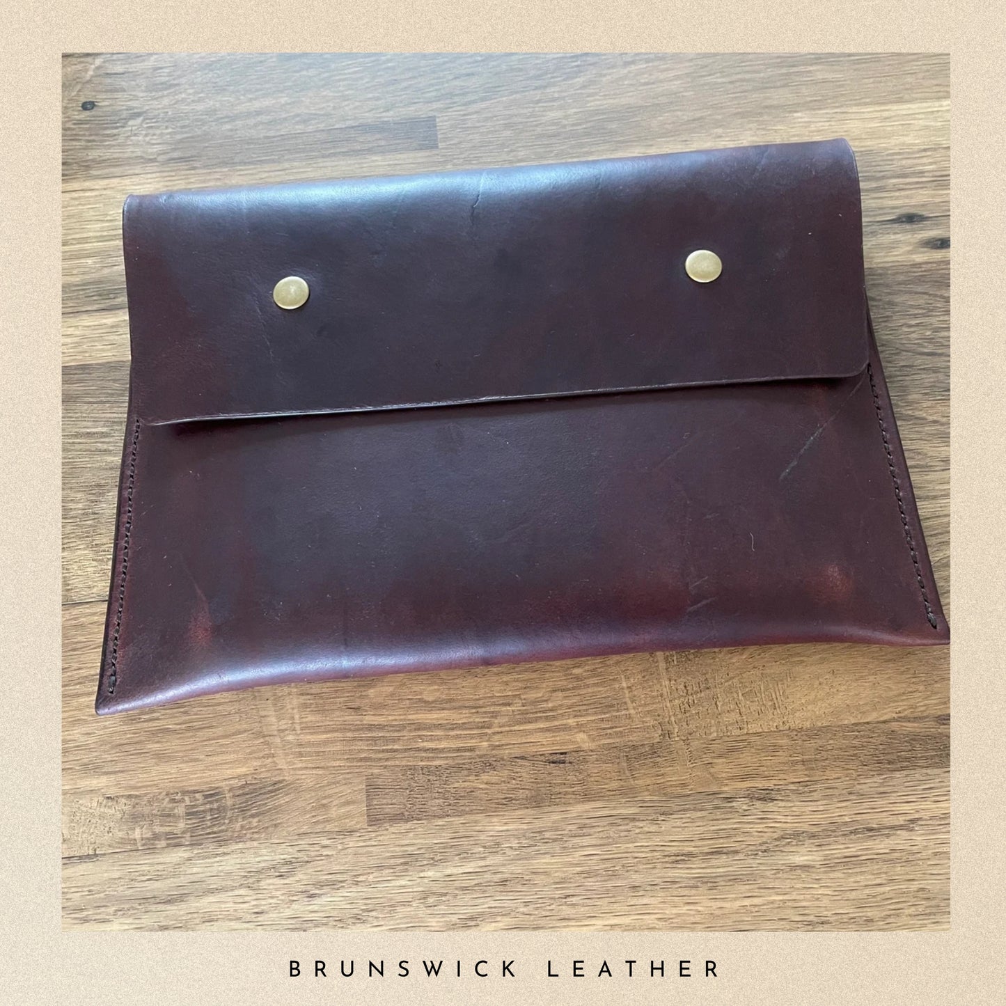 Brown leather clutch with gold studs on a wooden surface, branded 'Brunswick Leather'.