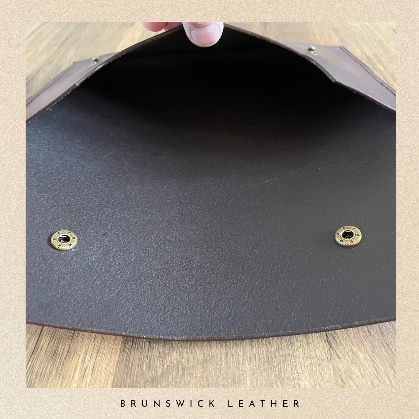 Close-up of a dark gray leather clutch with gold studs on a wooden surface, branded 'Brunswick Leather'.