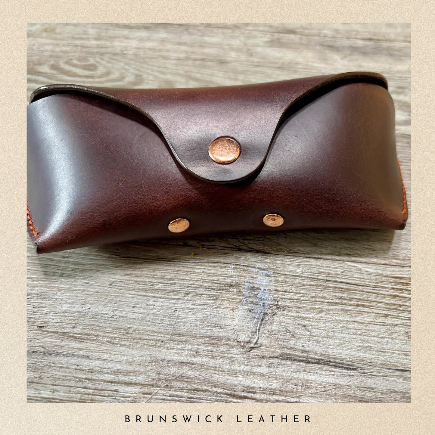 Brown leather case with gold studs on a wooden surface, branded 'Brunswick Leather'.