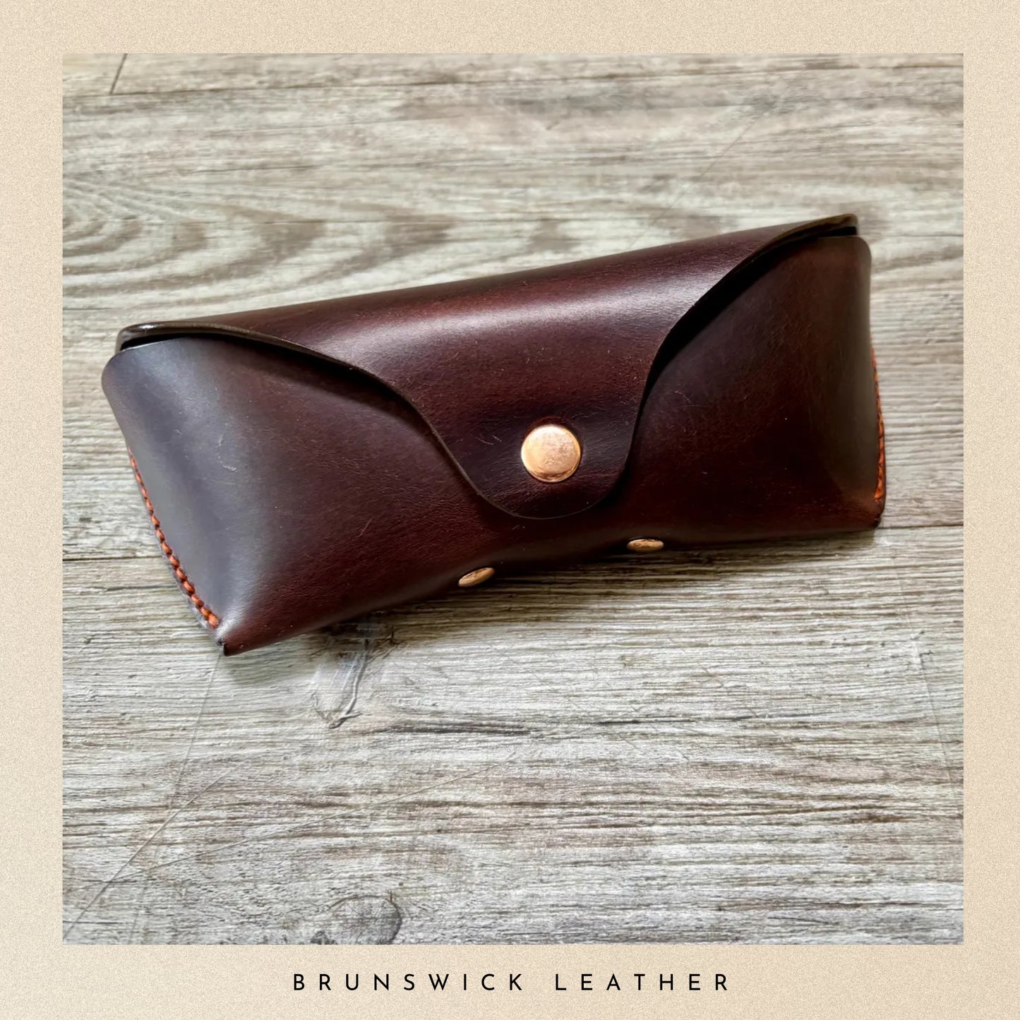 Brown leather case with a gold button on a wooden surface, branded 'Brunswick Leather'.