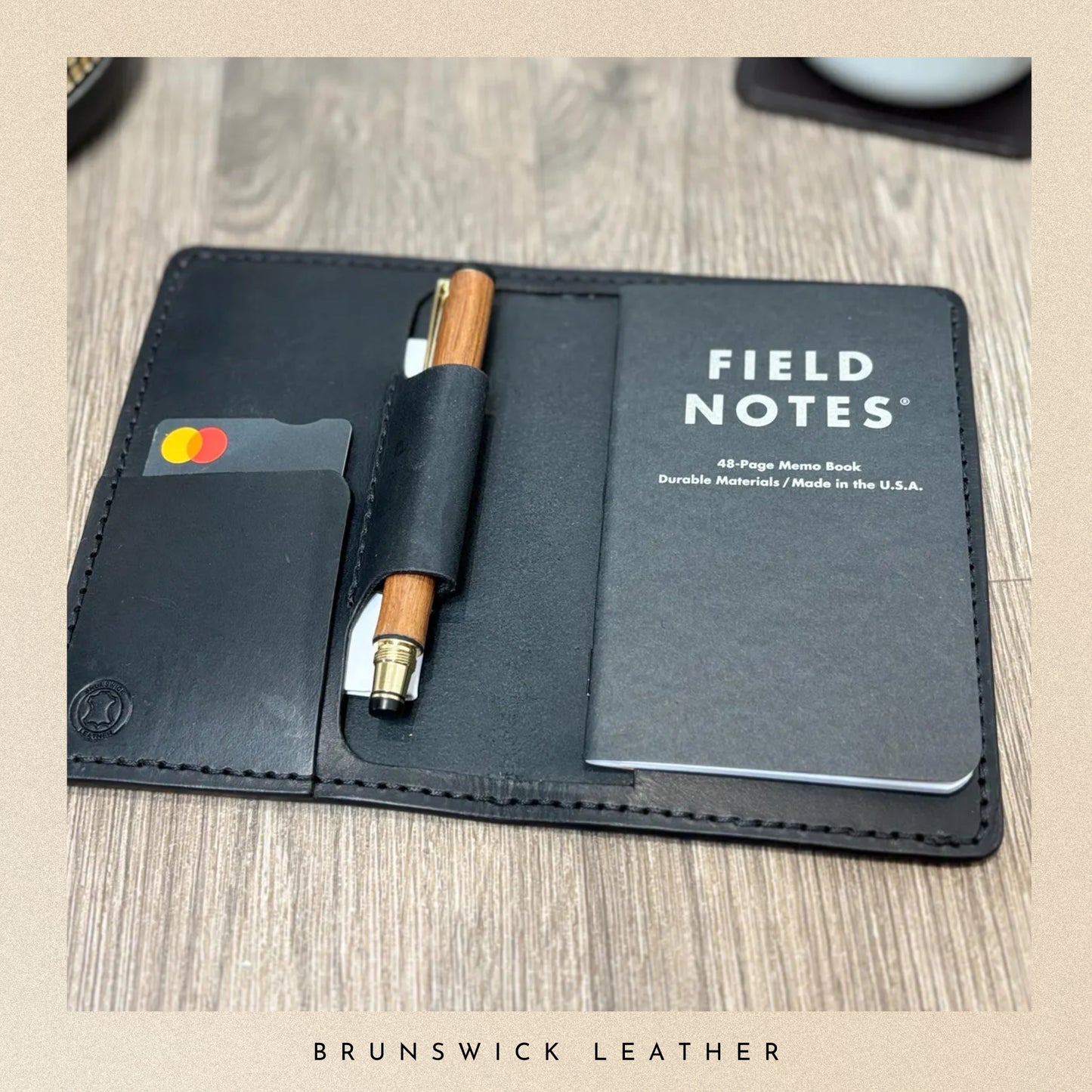 Black leather wallet with Field Notes notebook and pen on a wooden surface, branded 'Brunswick Leather'.