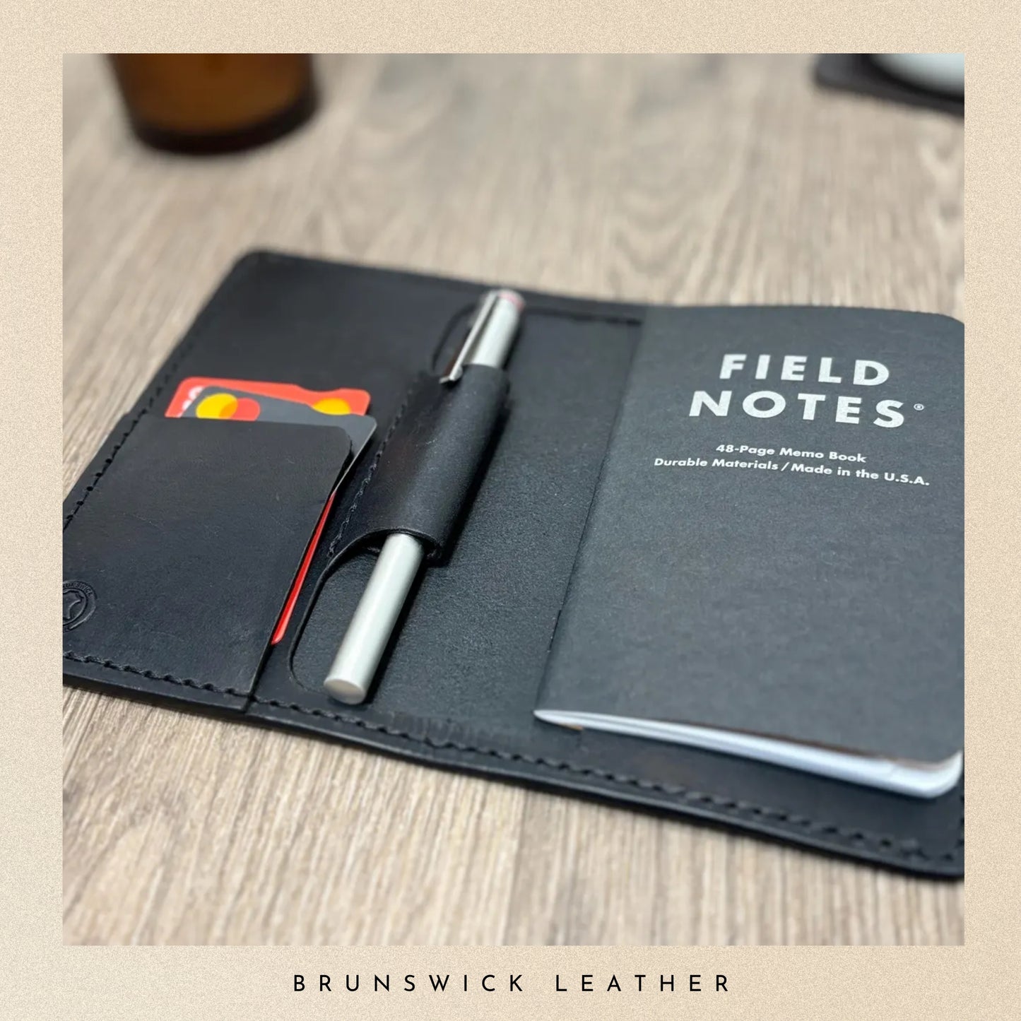 Field Notes notebook with a pen and card holder on a wooden surface, branded by Brunswick Leather.