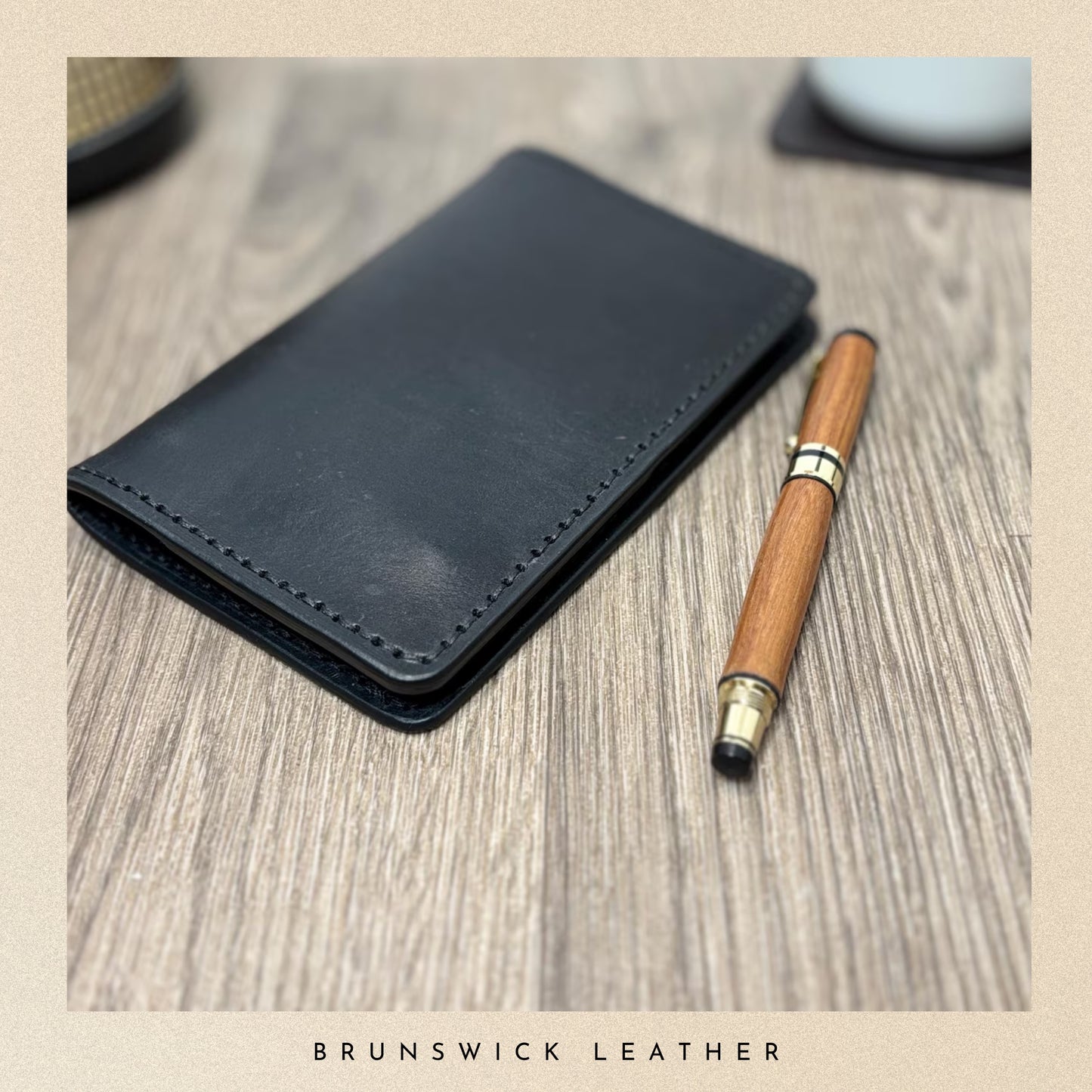Black leather wallet and wooden pen on a wooden surface with 'Brunswick Leather' branding.