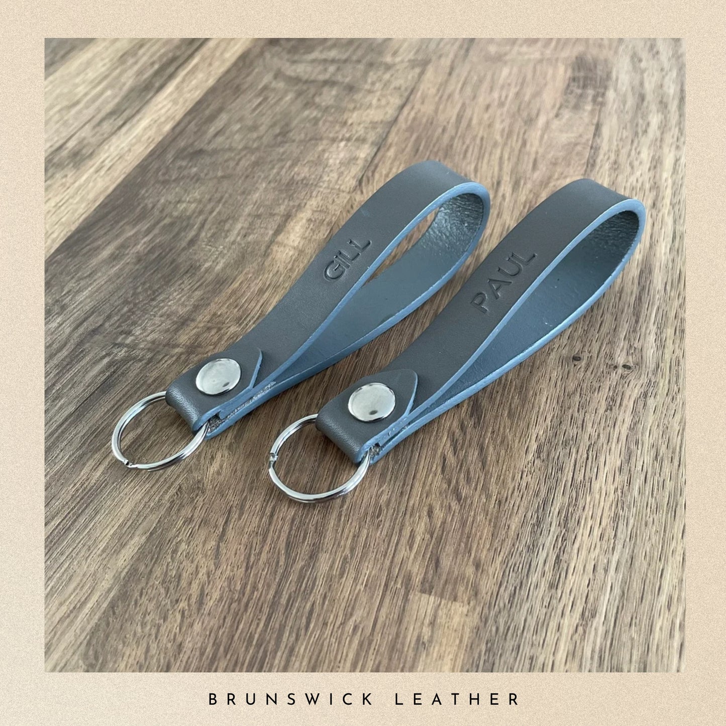 Two grey leather keychains on a wooden surface with 'Brunswick Leather' branding.